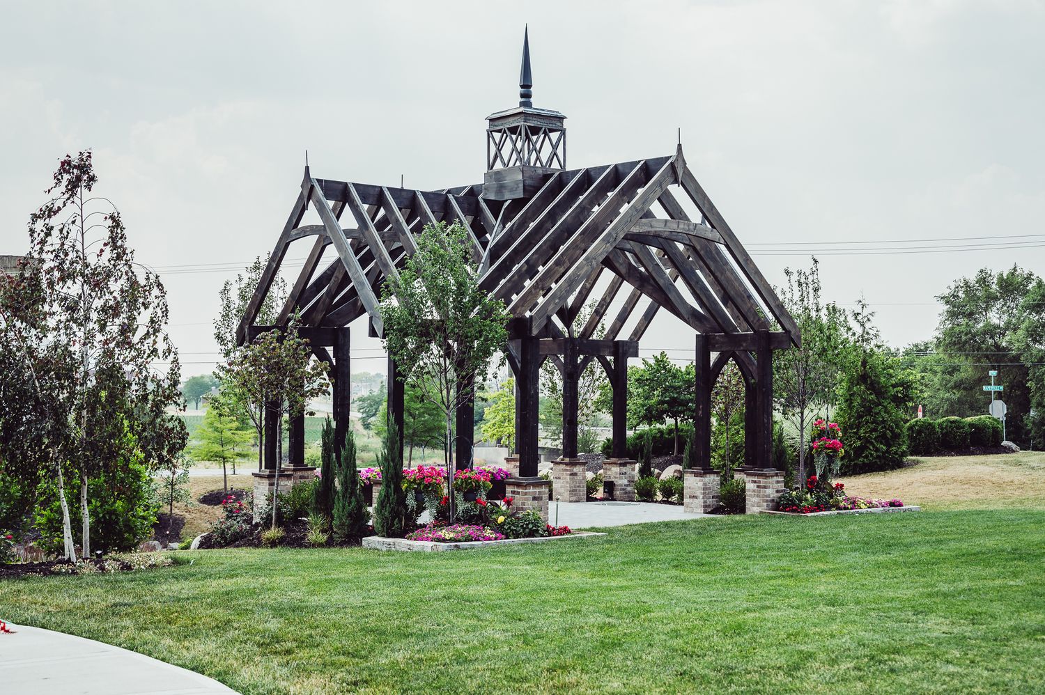 Elegant dark wood open-air pavilion with ornate spire surrounded by lush gardens and manicured lawn.