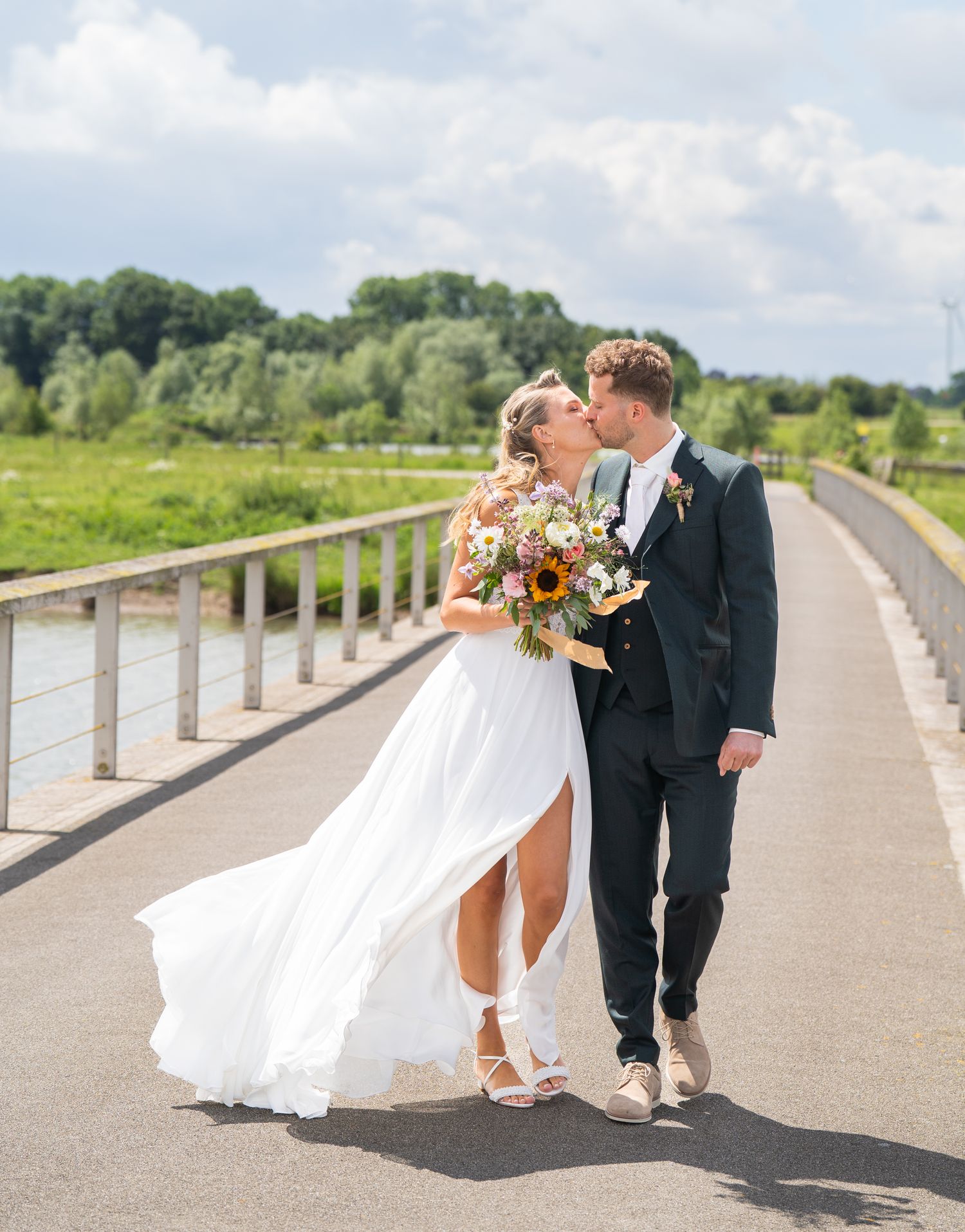 Bride and groom kissing on a sunny bridge, bride holding sunflower bouquet in flowing white wedding dress.