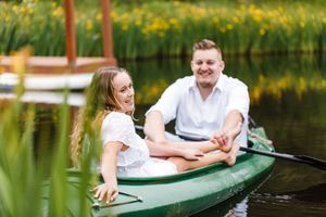 Couple relaxing in a green canoe surrounded by yellow wildflowers along a tranquil lake shore.
