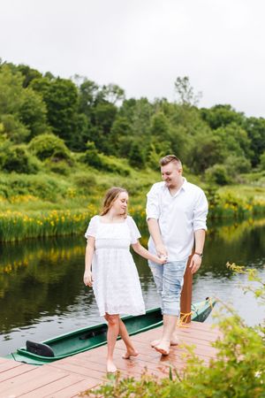Couple in white outfits standing on wooden dock near a green canoe on a summer lake.