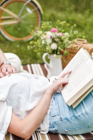 Person relaxing in grass while reading a book on a sunny day with flowers nearby.