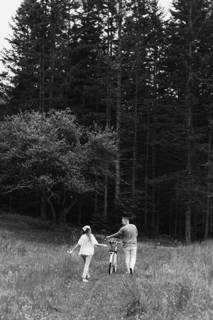 Black and white image of people walking with a bicycle through a meadow bordered by pine trees.