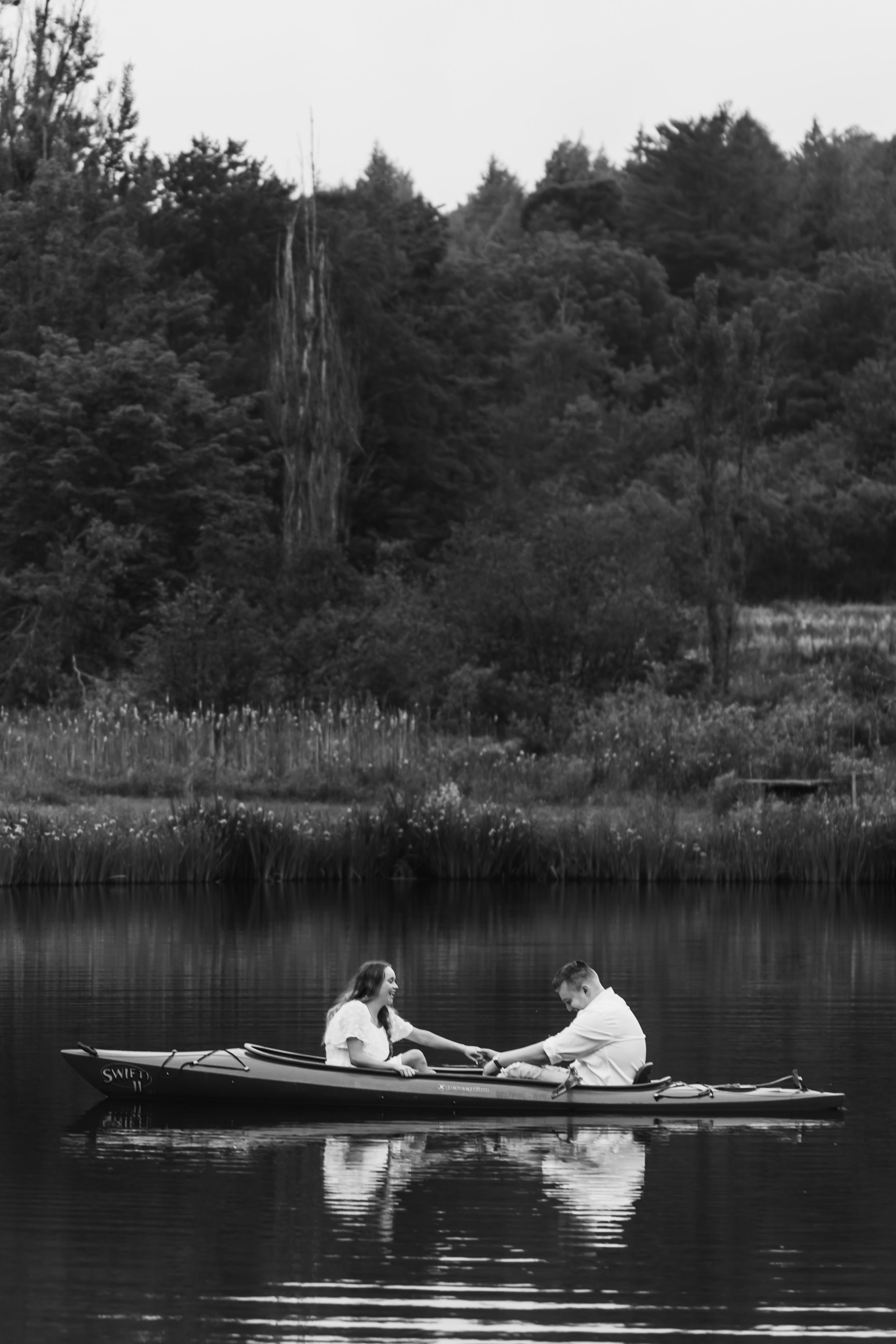 Two people rowing in a wooden boat on a tranquil lake surrounded by tall reeds and forest in black and white.