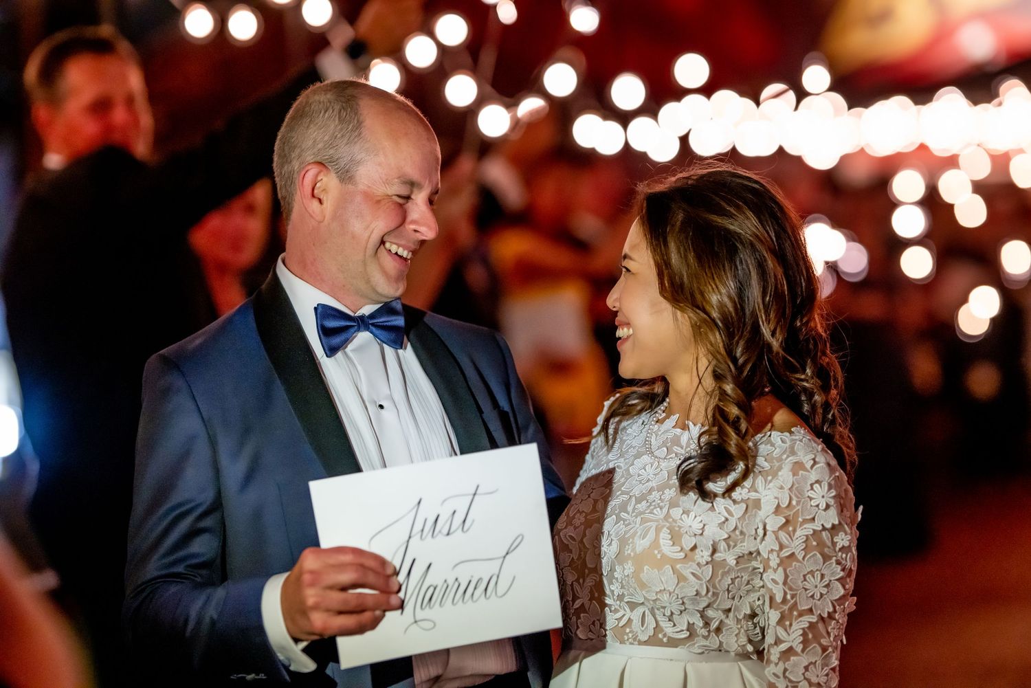 Newlywed couple smiling at each other holding a Just Married sign at a romantic wedding reception.