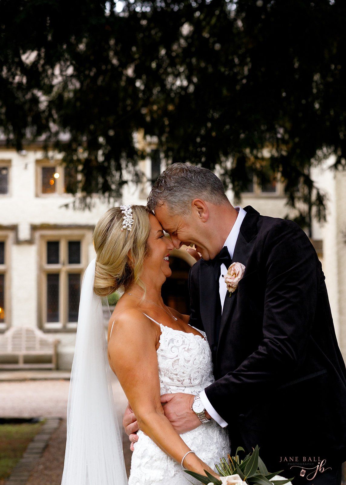 Bride and groom share a romantic forehead touch in wedding attire outside an elegant manor. Mitton Hall