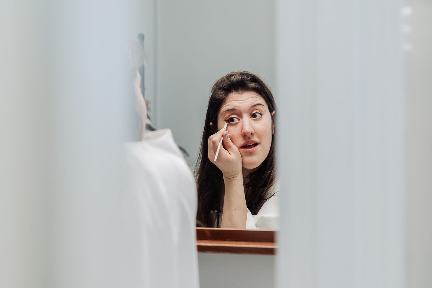 Woman applying eye makeup while looking in a bathroom mirror, beauty routine at home.