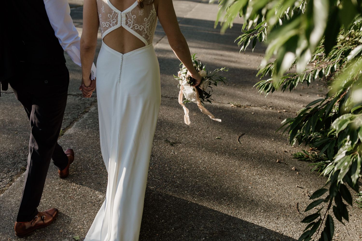 Back view of an elegant white wedding dress as couple walks down a garden path.