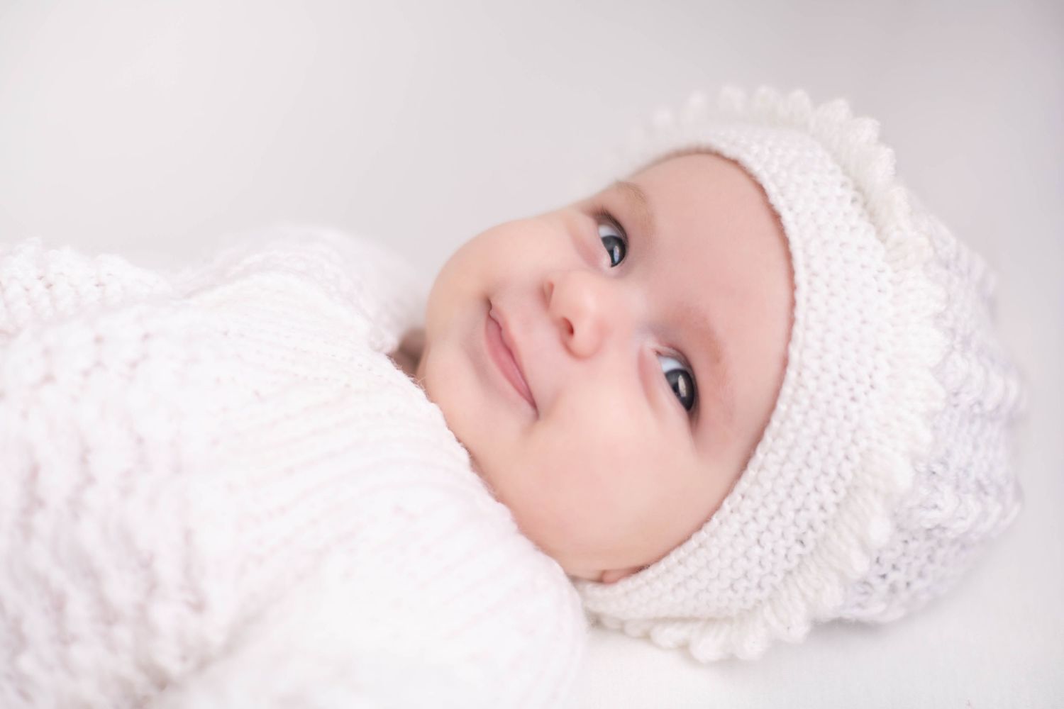 Adorable smiling baby wearing a white knit hat and soft white blanket wrap, with bright blue eyes.
