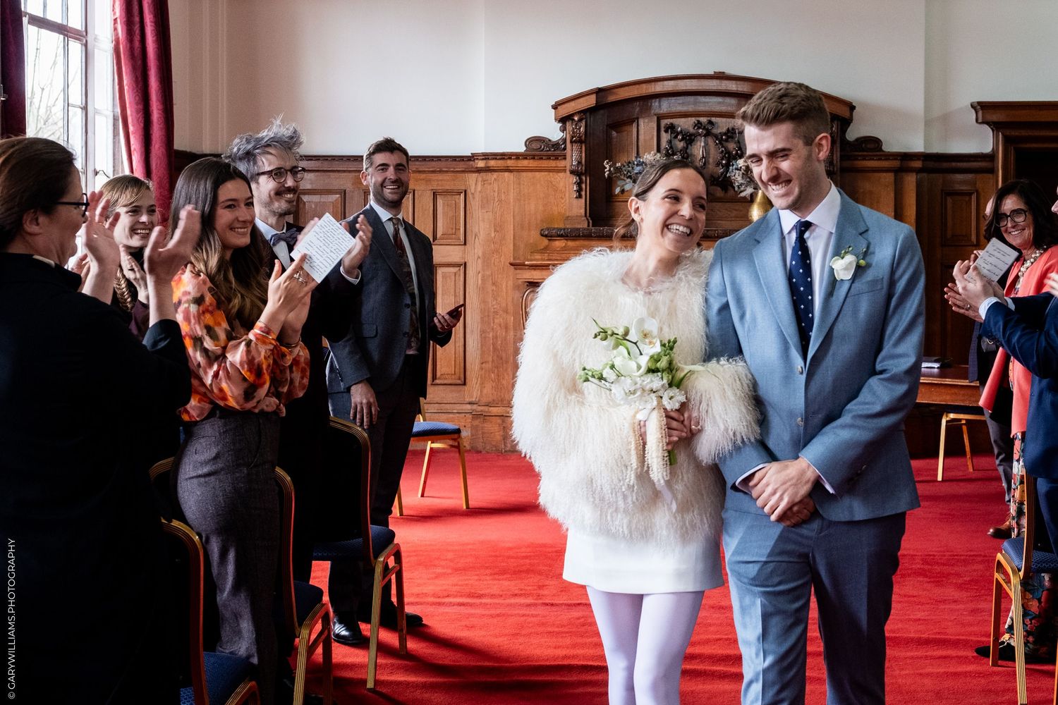 A bride and groom stand together on a red carpet, surrounded by their wedding party and guests.