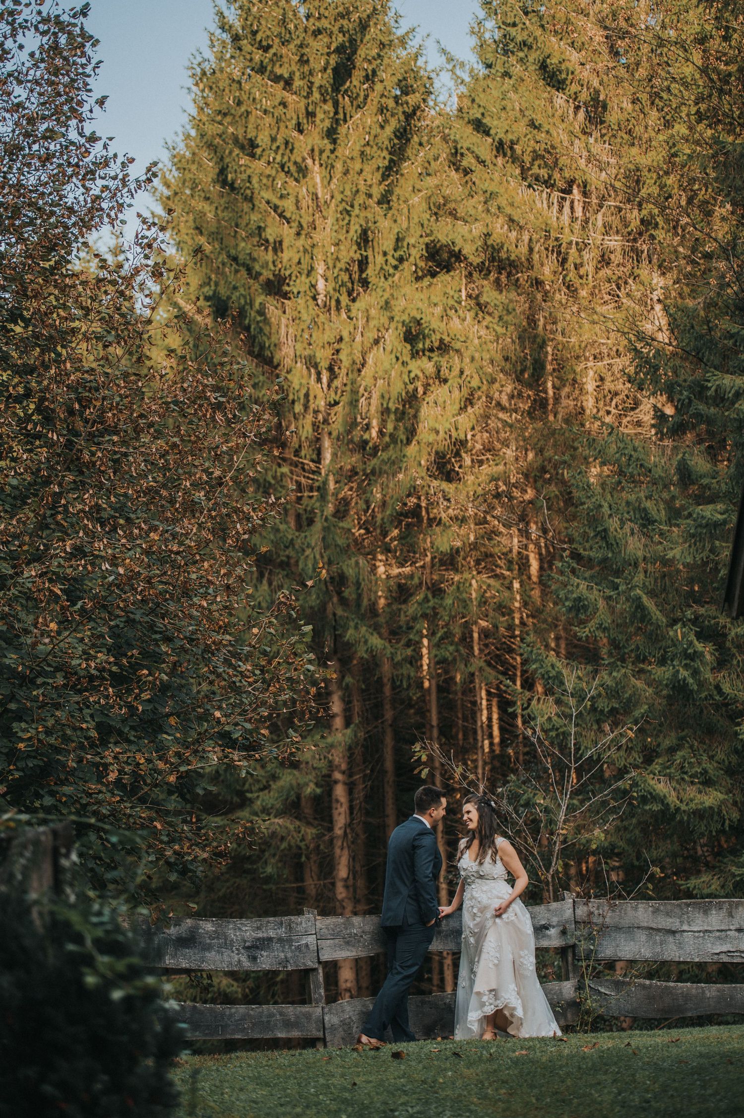 Romantic couple stands beside rustic wooden fence at sunset with tall evergreen trees creating a natural backdrop.
