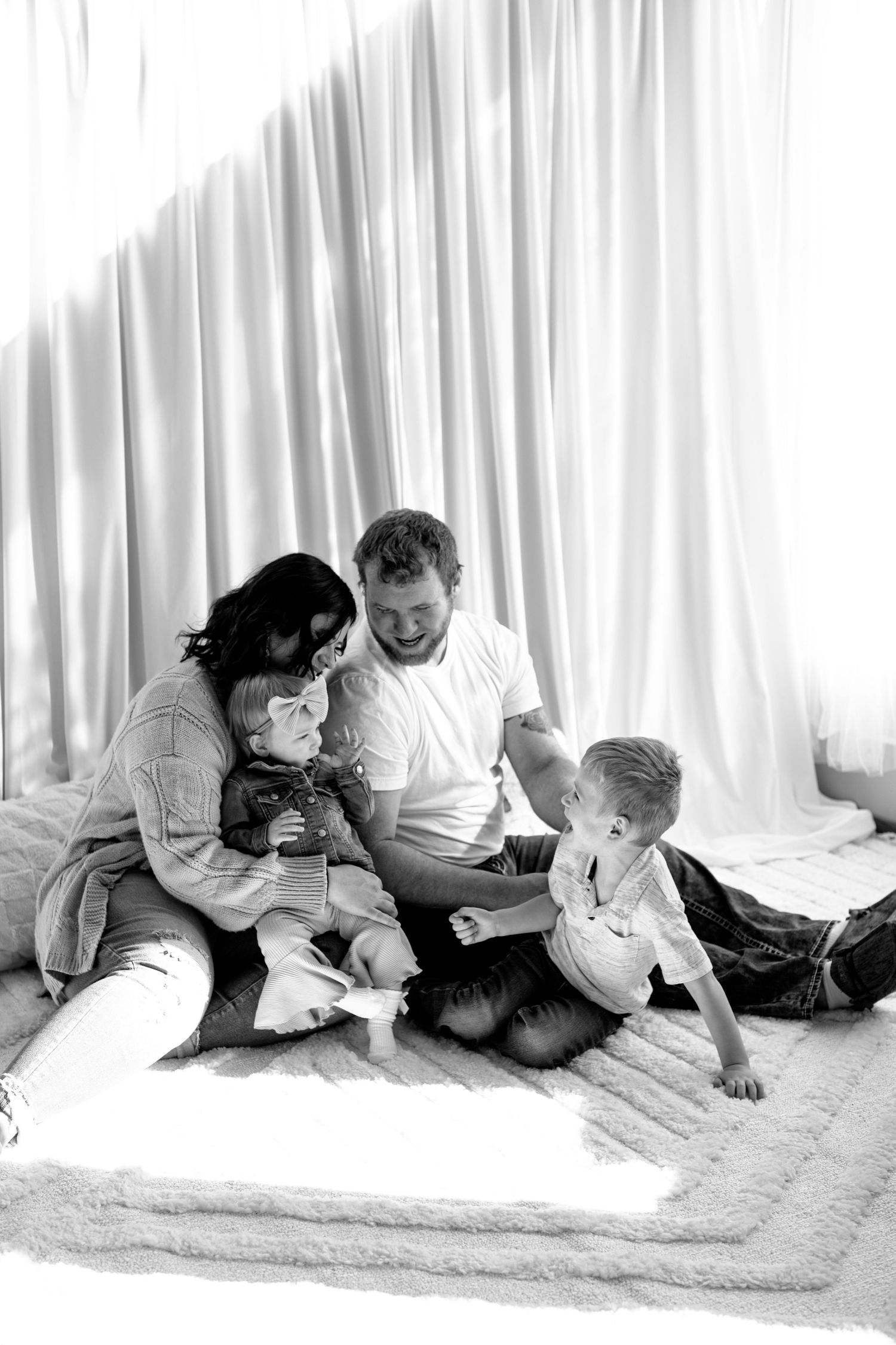 Family lounging together on white carpet in front of sheer curtains in black and white photo series.