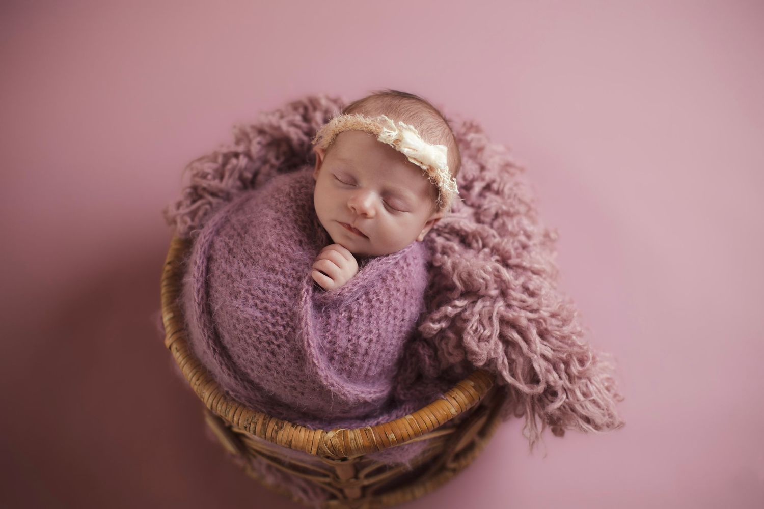 Sleeping newborn in purple knit wrap nestled in woven basket.