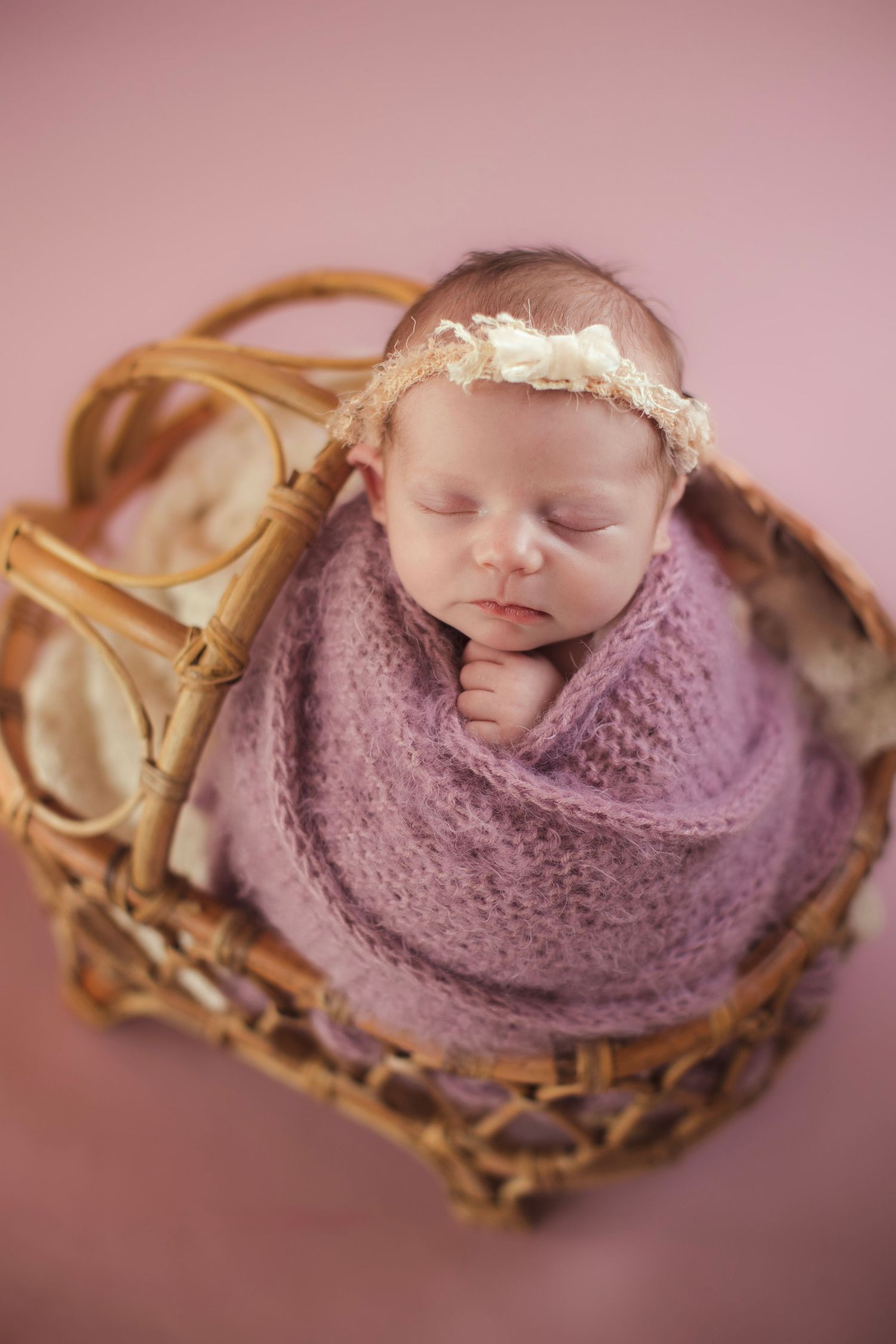 Newborn baby wrapped in purple knit blanket sleeping in rustic wooden basket.