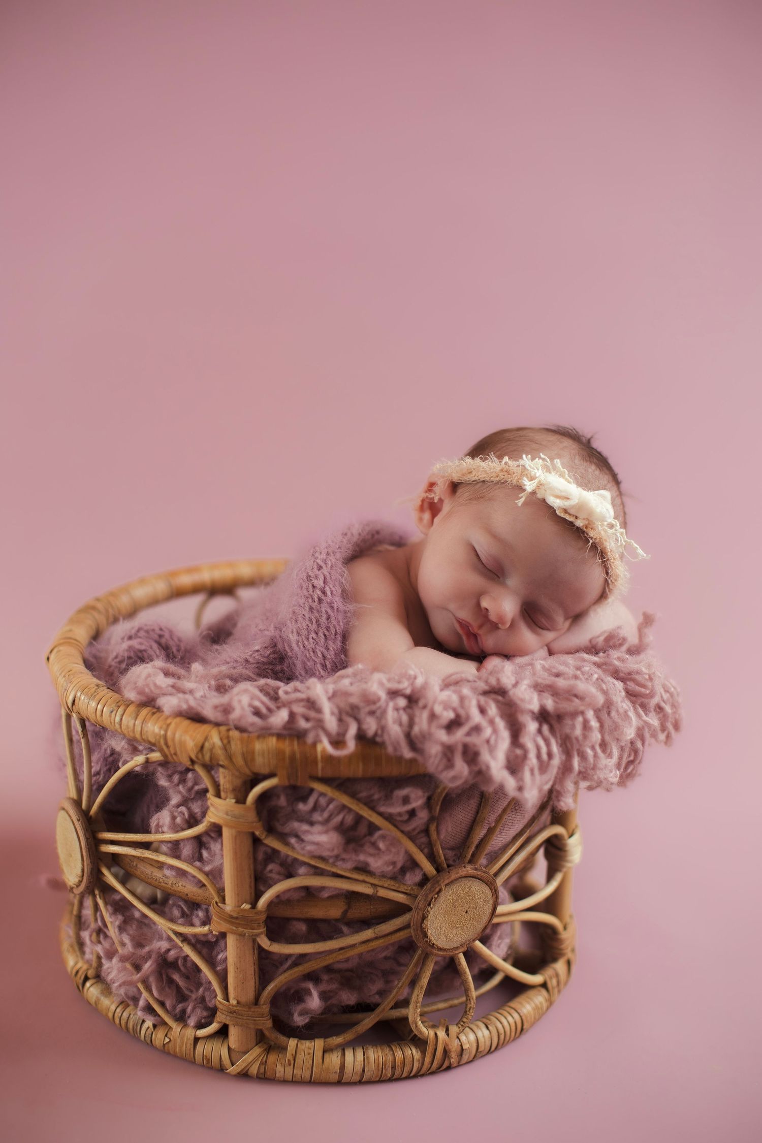 Newborn baby sleeping peacefully in a woven basket with pink textured blanket against rose background.