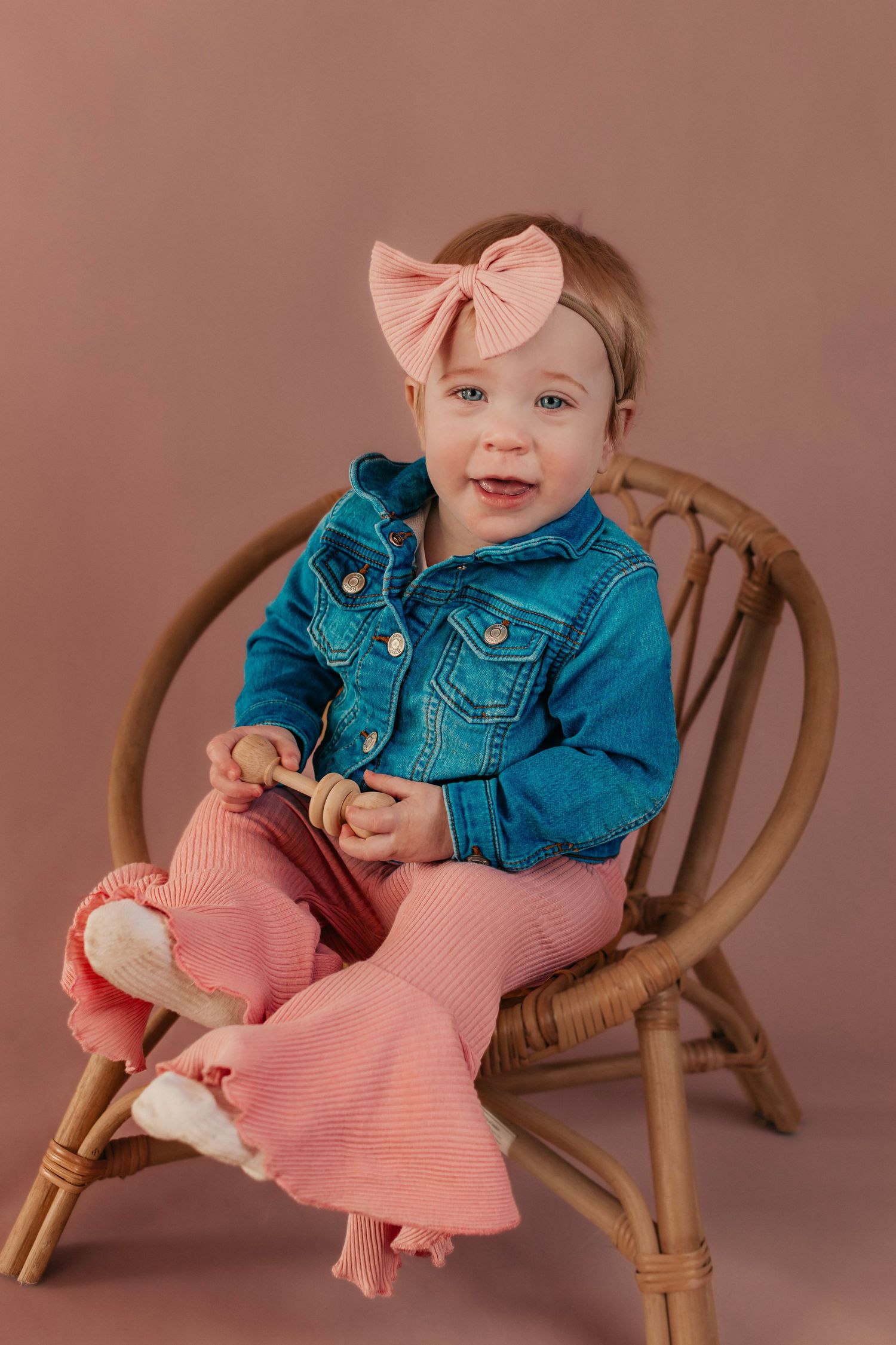 Baby sitting in rattan chair wearing teal denim jacket and pink dress with matching bow.