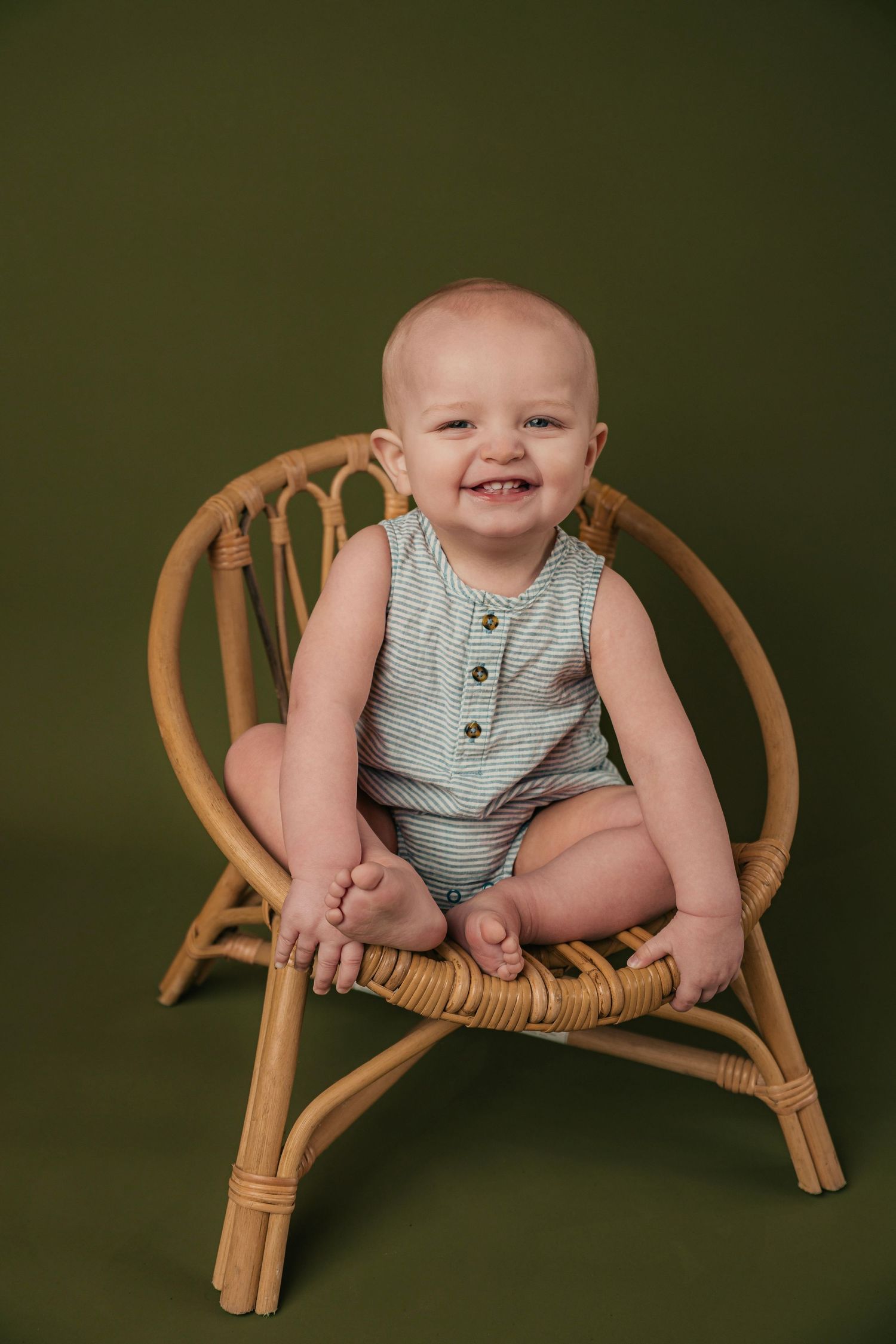 Baby in striped romper sitting in wooden chair against dark green backdrop.