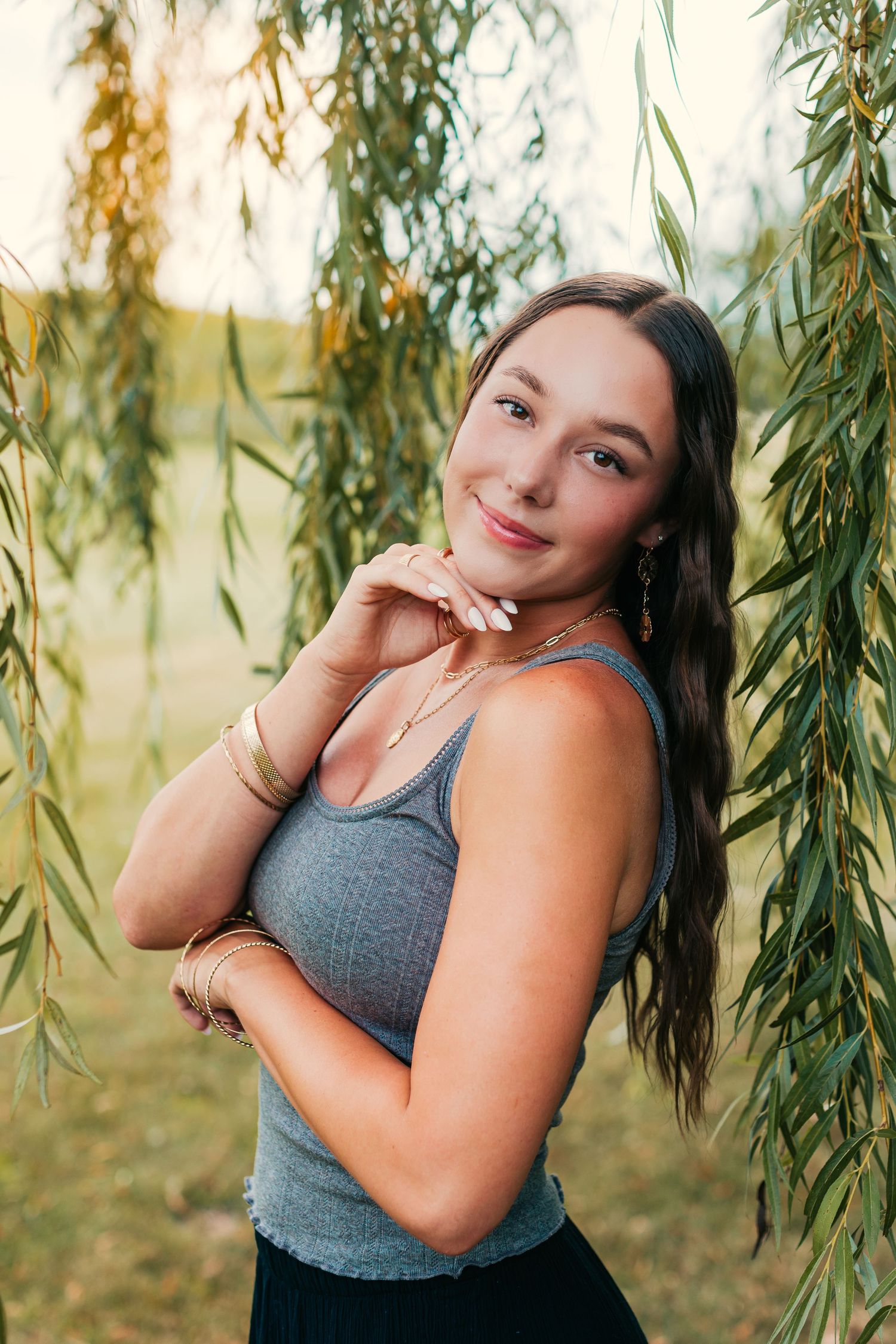 Young woman in gray tank top poses playfully under willow tree branches in natural lighting.