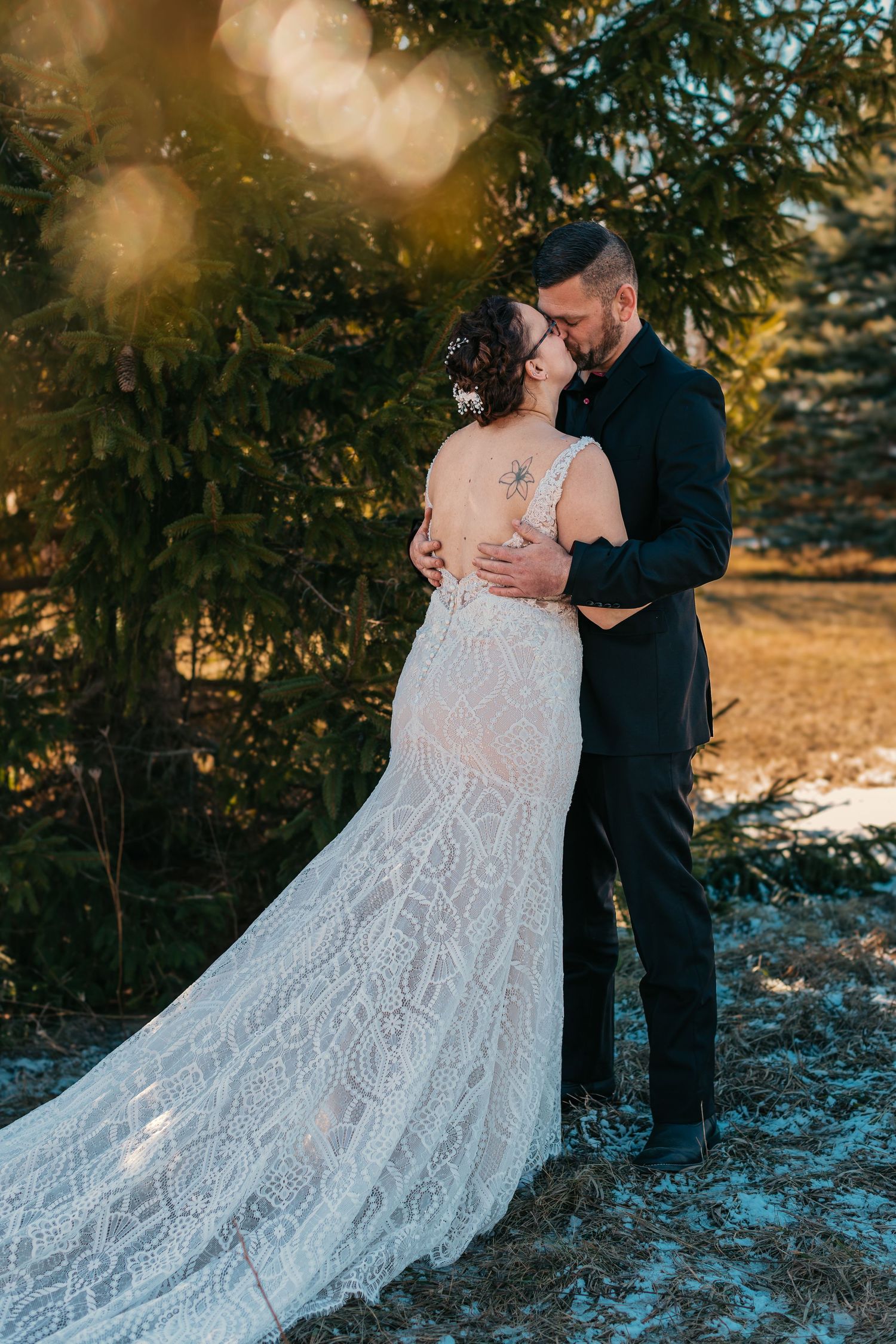 Couple in wedding attire embracing outdoors with sunlit evergreen trees in background.
