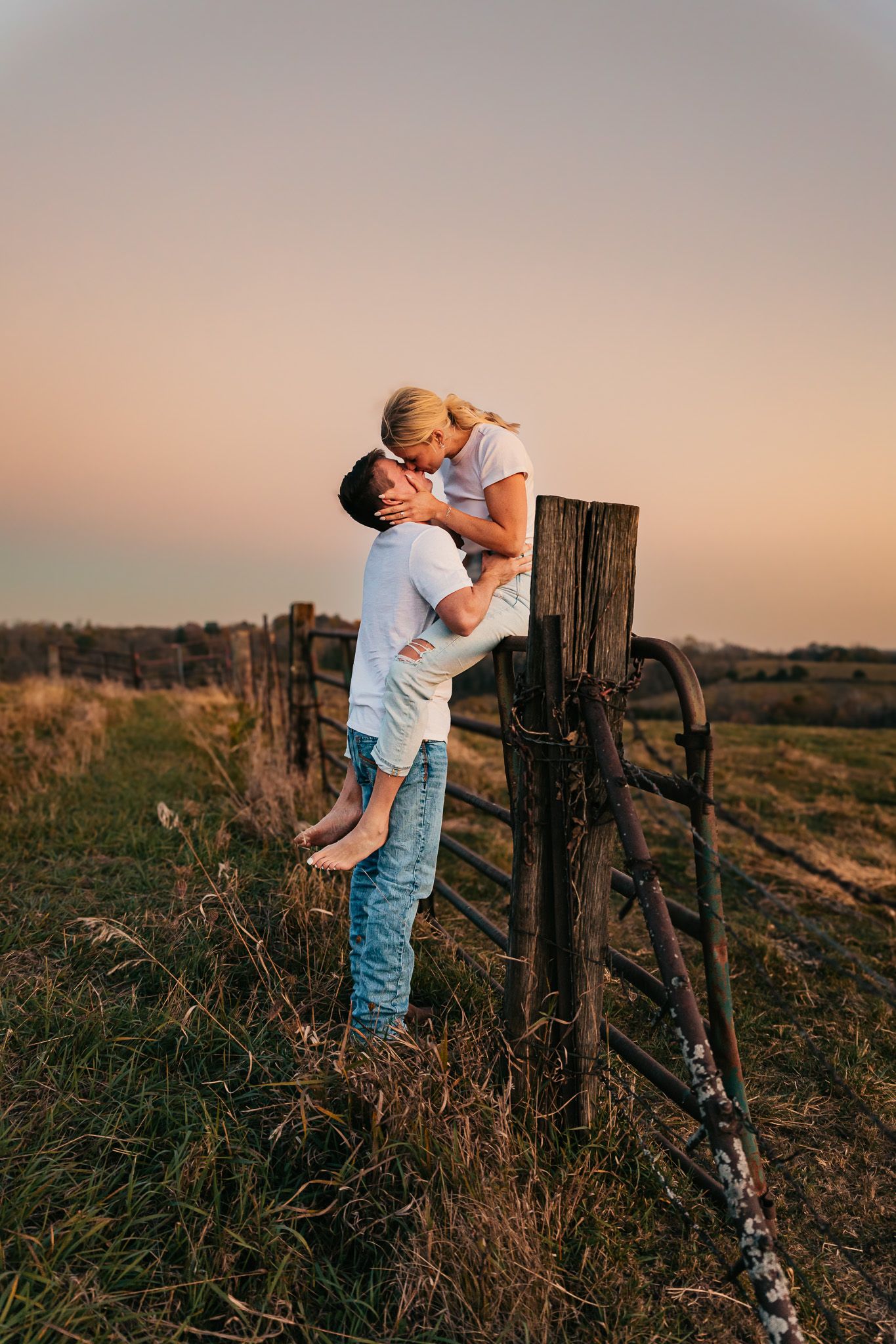 Couple embracing by rustic wooden fence post during golden hour sunset.