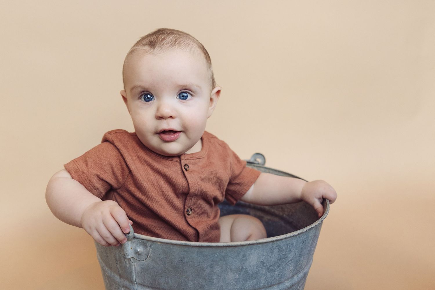 Baby wearing brown onesie sits in rustic metal bucket against neutral backdrop.