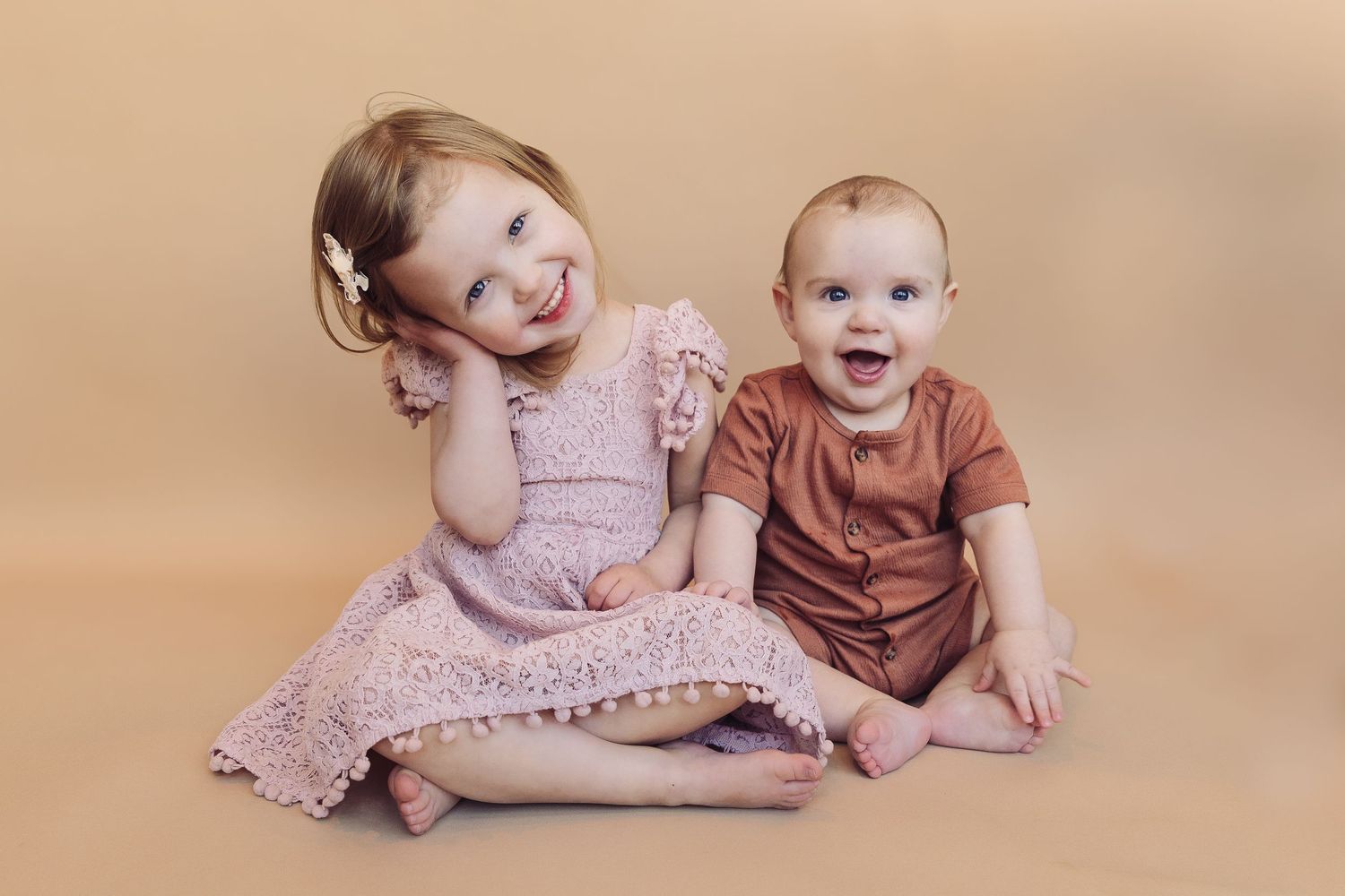 Two young children sitting together on beige backdrop wearing pink and brown outfits.