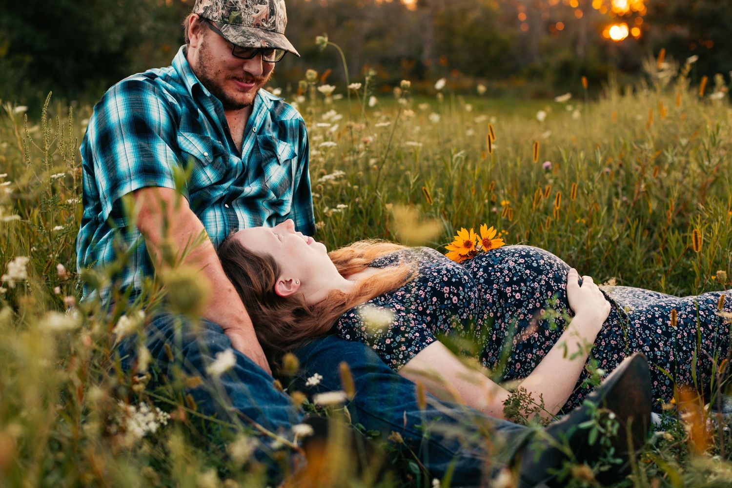 Couple lying in tall grass with sunflowers at sunset during a romantic photoshoot.