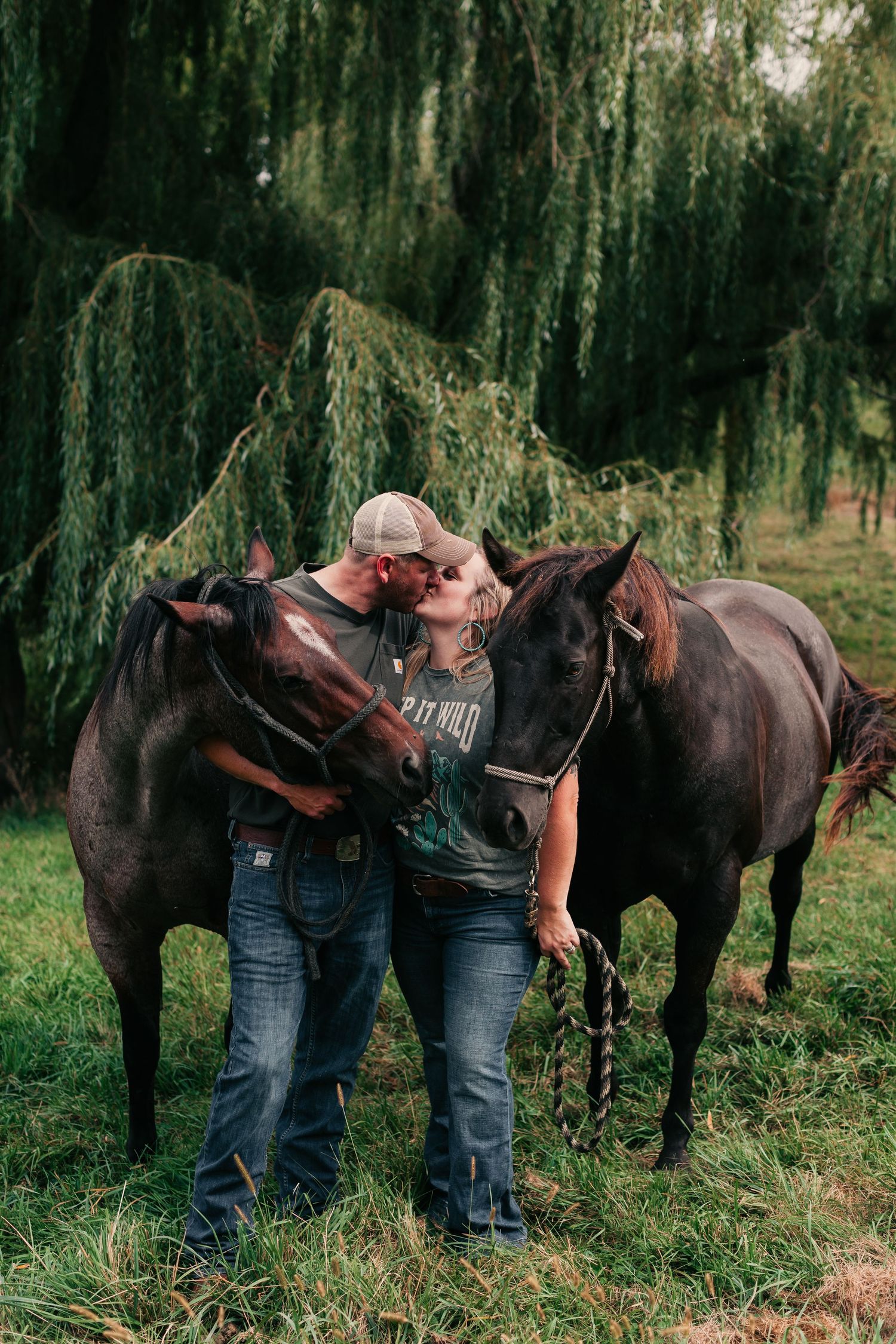 Two people in casual attire standing between two horses near a weeping willow tree.