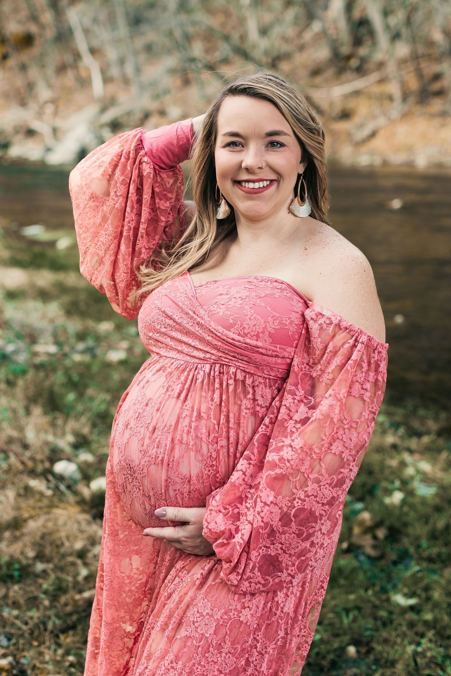 Pregnant woman in coral lace dress posing outdoors by water.