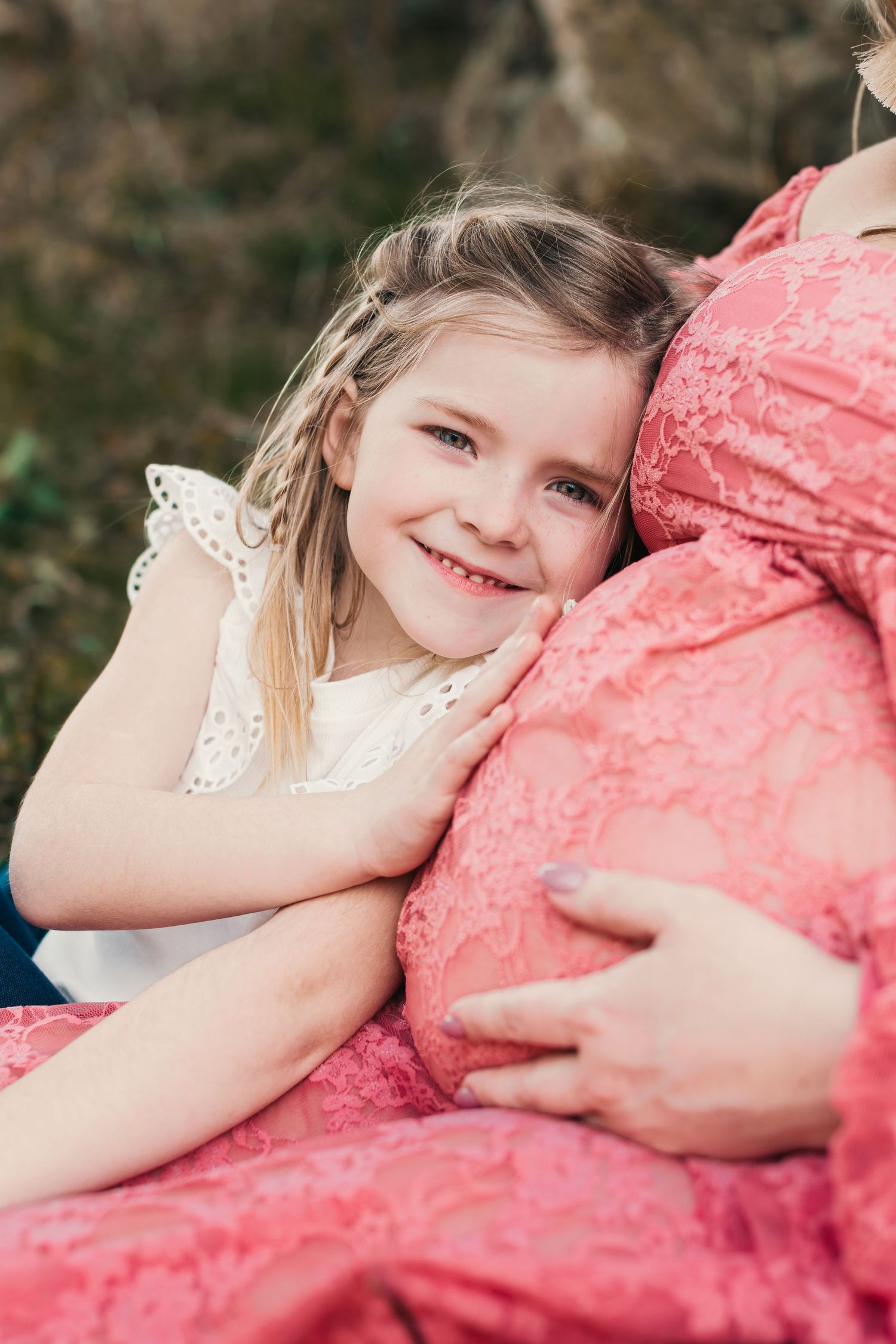 Young child in white dress leaning against pink fabric outdoors.