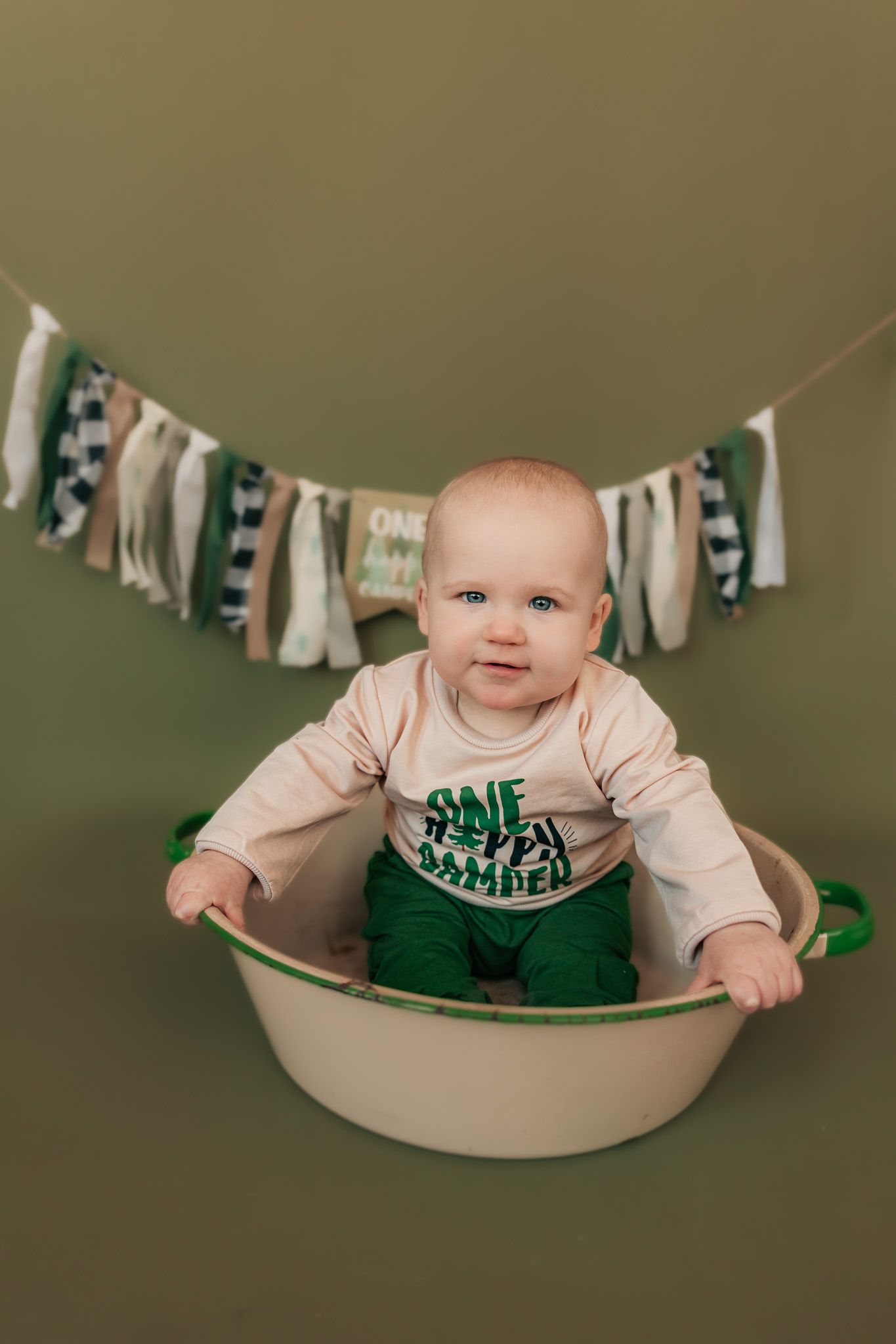 Baby wearing green outfit sitting with festive banner decorations.