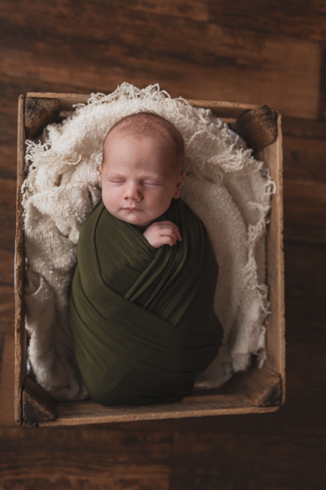 Sleeping newborn wrapped in olive green swaddle rests in wooden box lined with white lace fabric.