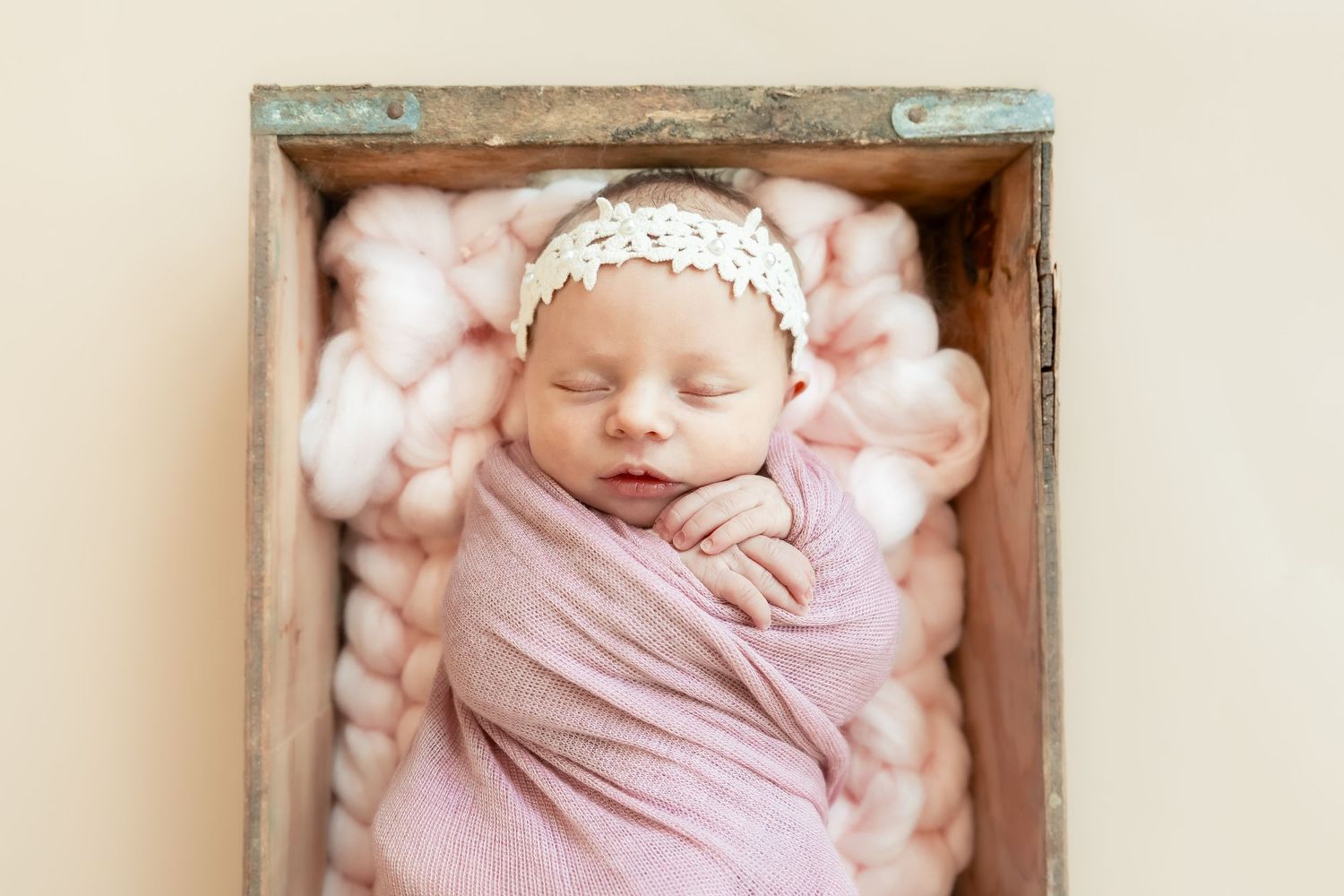 Newborn baby sleeping peacefully in wooden box with pink wrap and lace details.