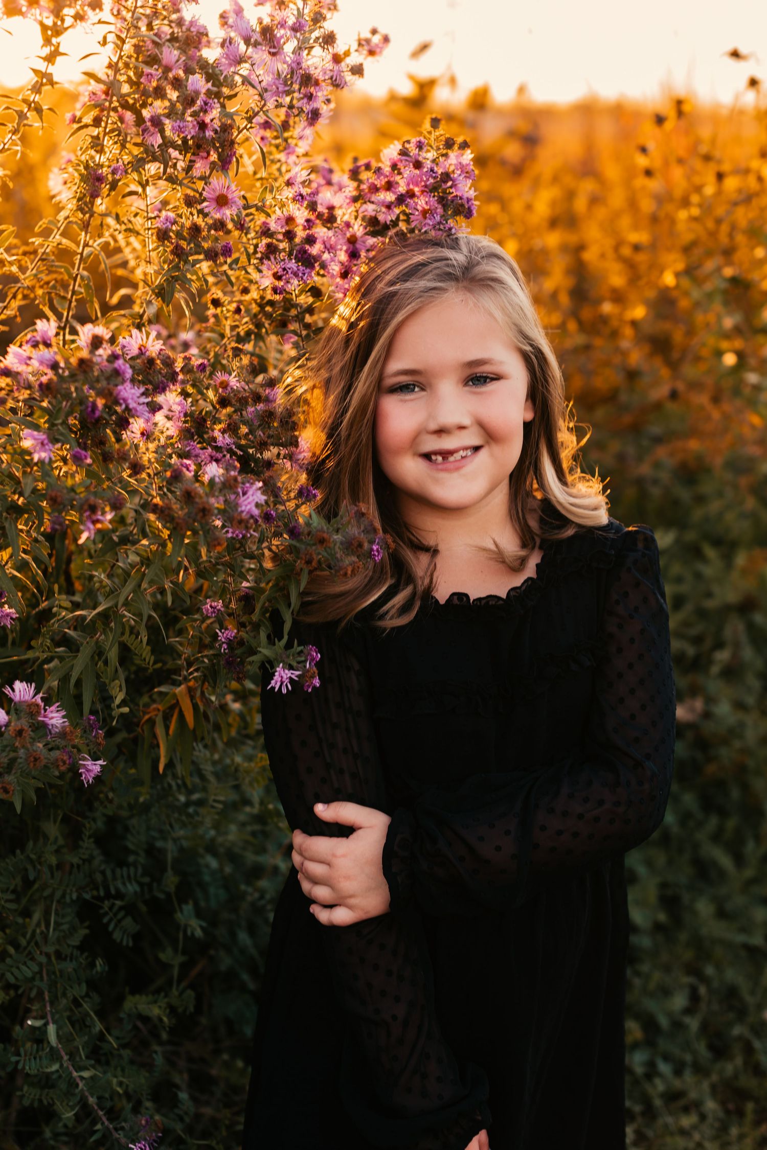 Young person in black dress standing next to purple flowers during golden hour.