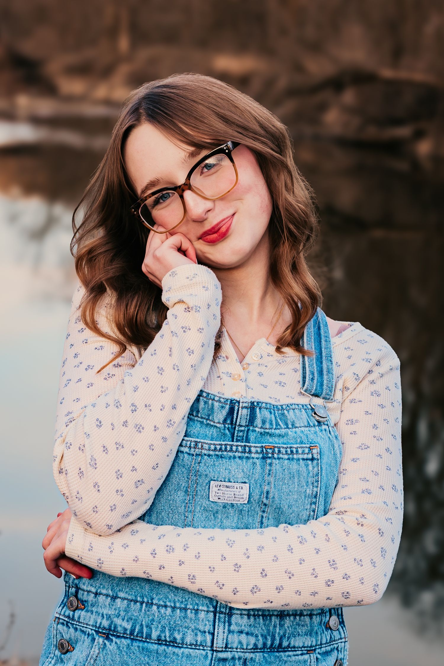 Casual portrait against lake backdrop wearing denim overalls and floral print blouse.