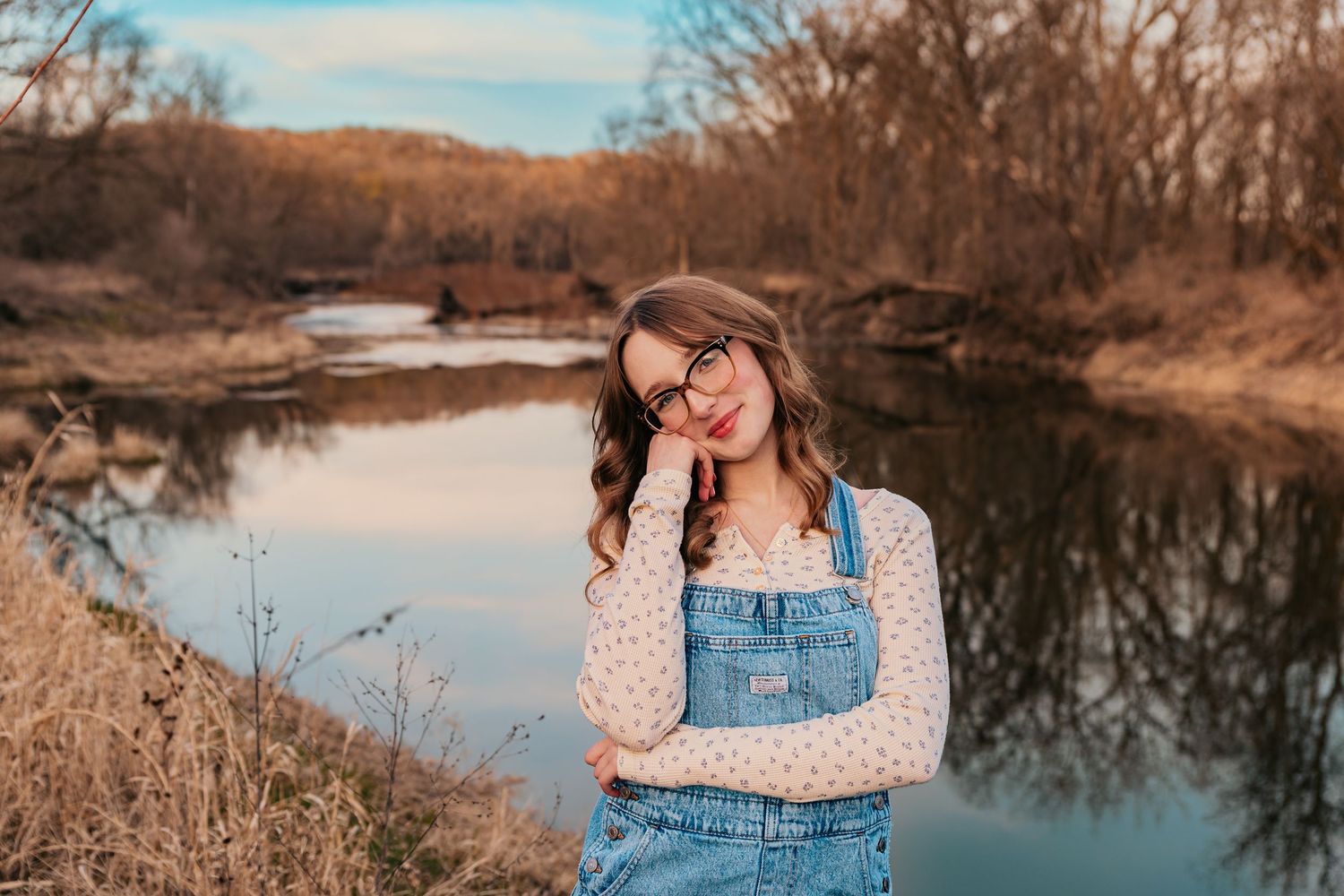 Outdoor portrait session at sunset by a reflective lake with denim overalls and floral top.