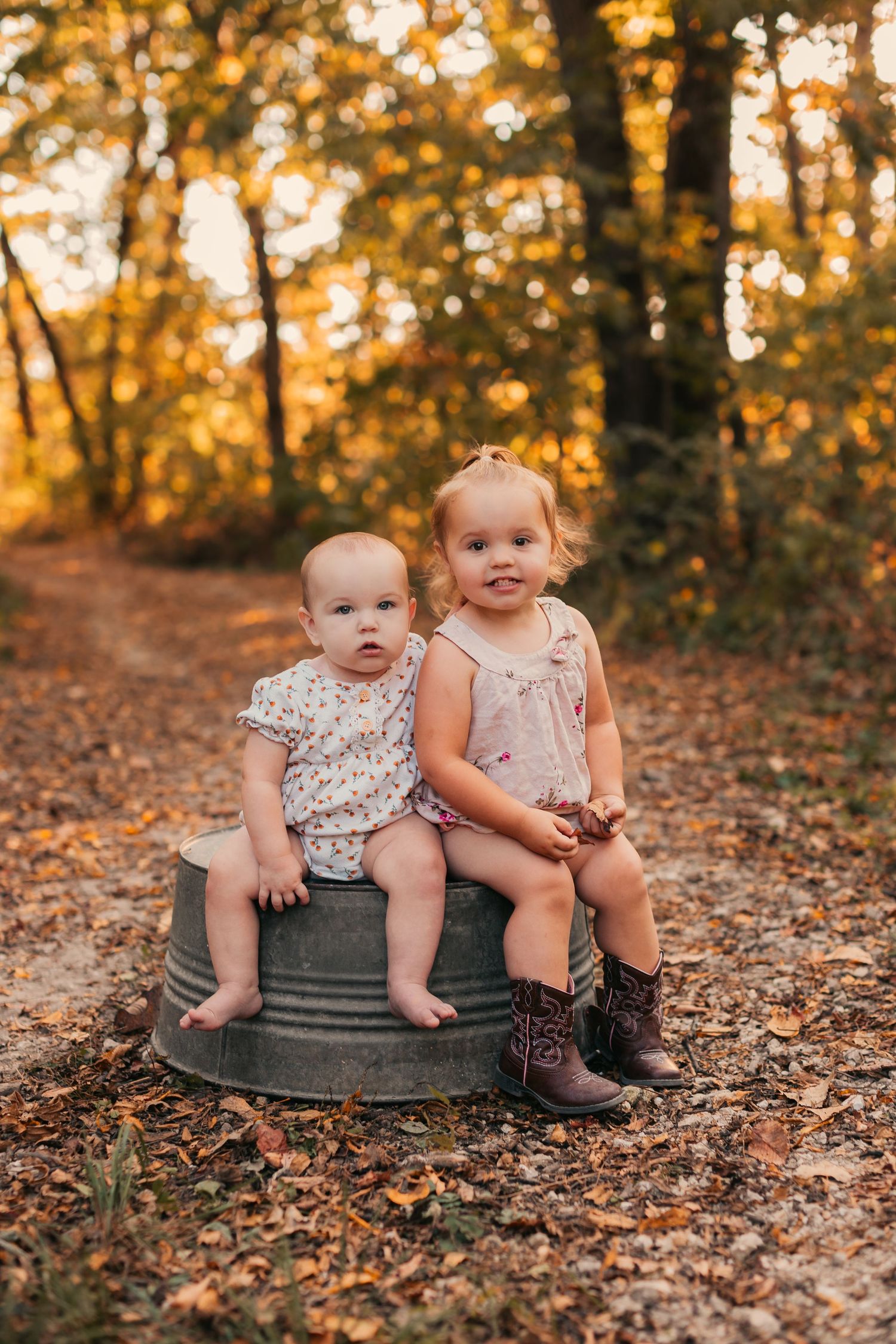 Children sit together on a metal tub surrounded by autumn leaves in a golden-lit forest setting.