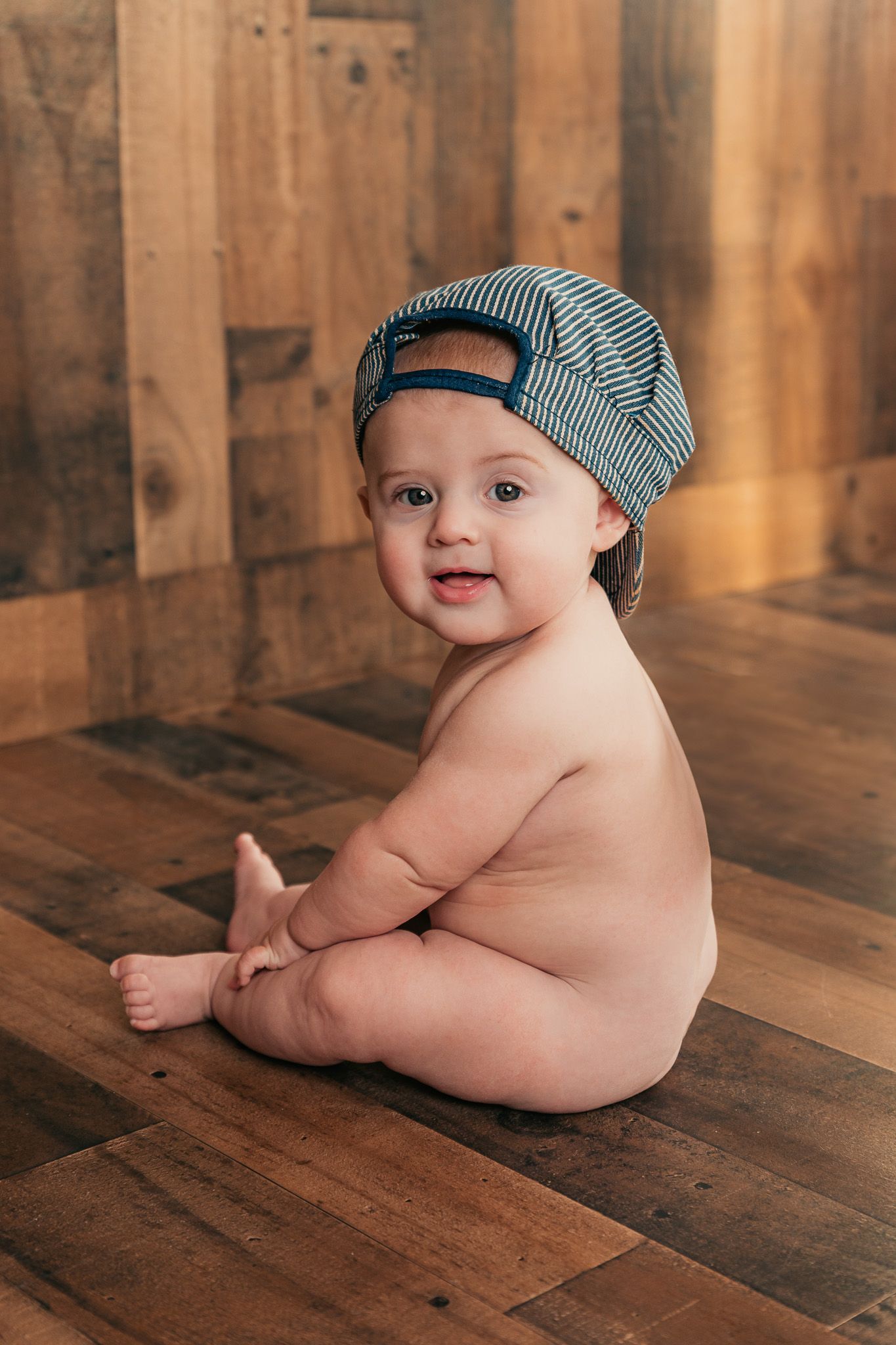 Baby wearing striped beanie poses on wooden floor during lifestyle photoshoot.