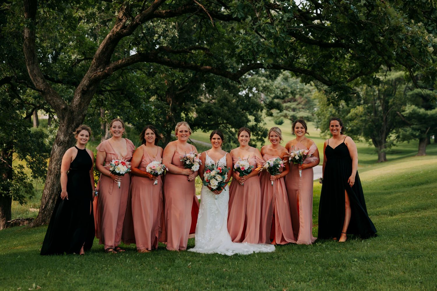 Wedding party poses in pink bridesmaid dresses and white gown outdoors in a park setting.