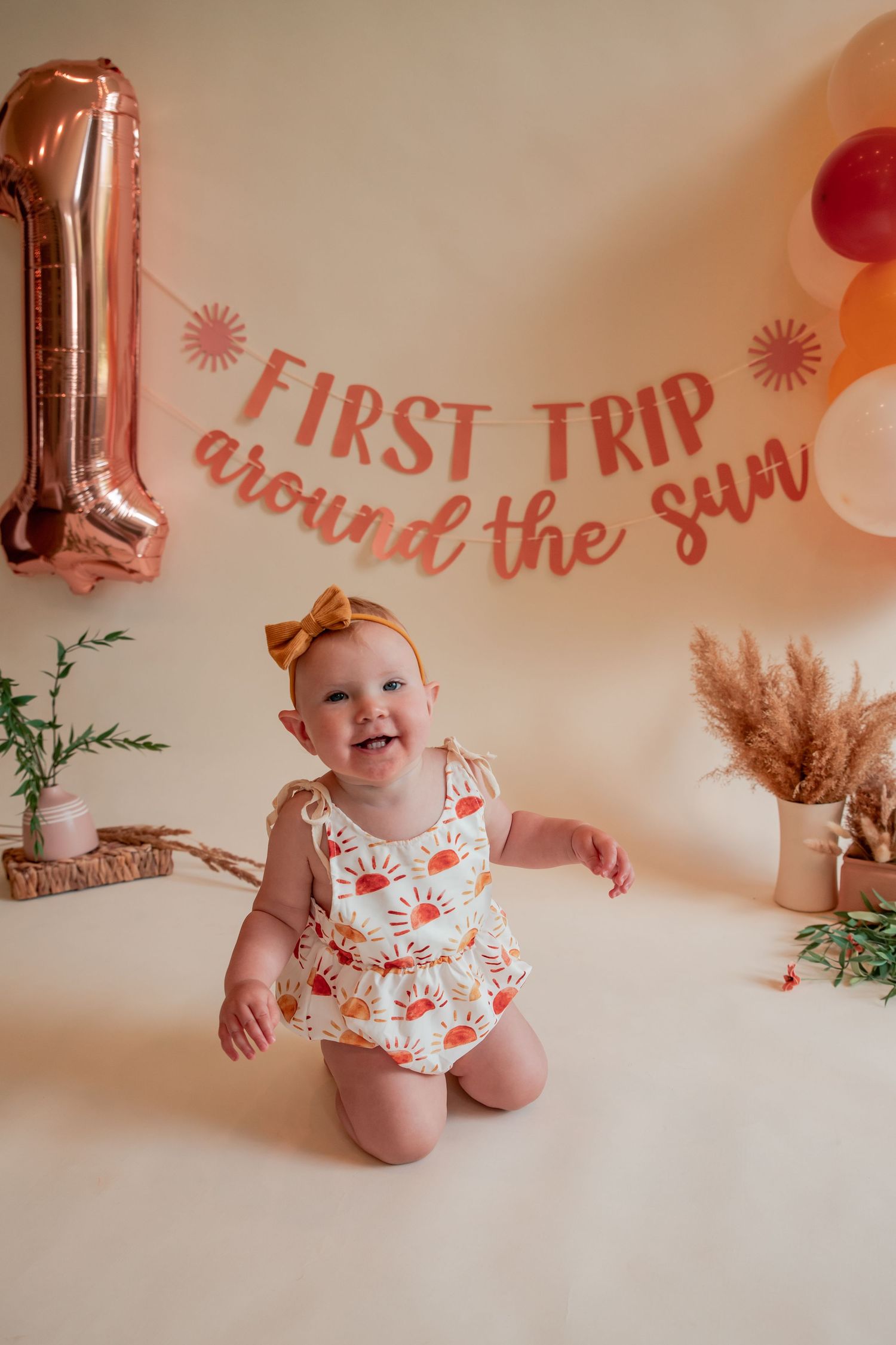 A baby celebrates their first birthday with a 'First Trip Around the Sun' banner and floral decorations.