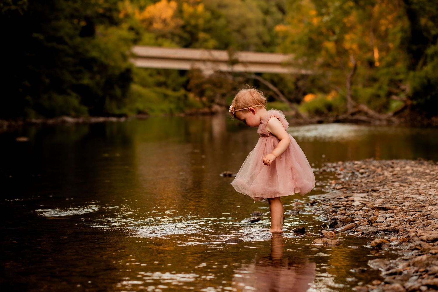 A child in a pink dress plays along a scenic riverbank during golden hour.