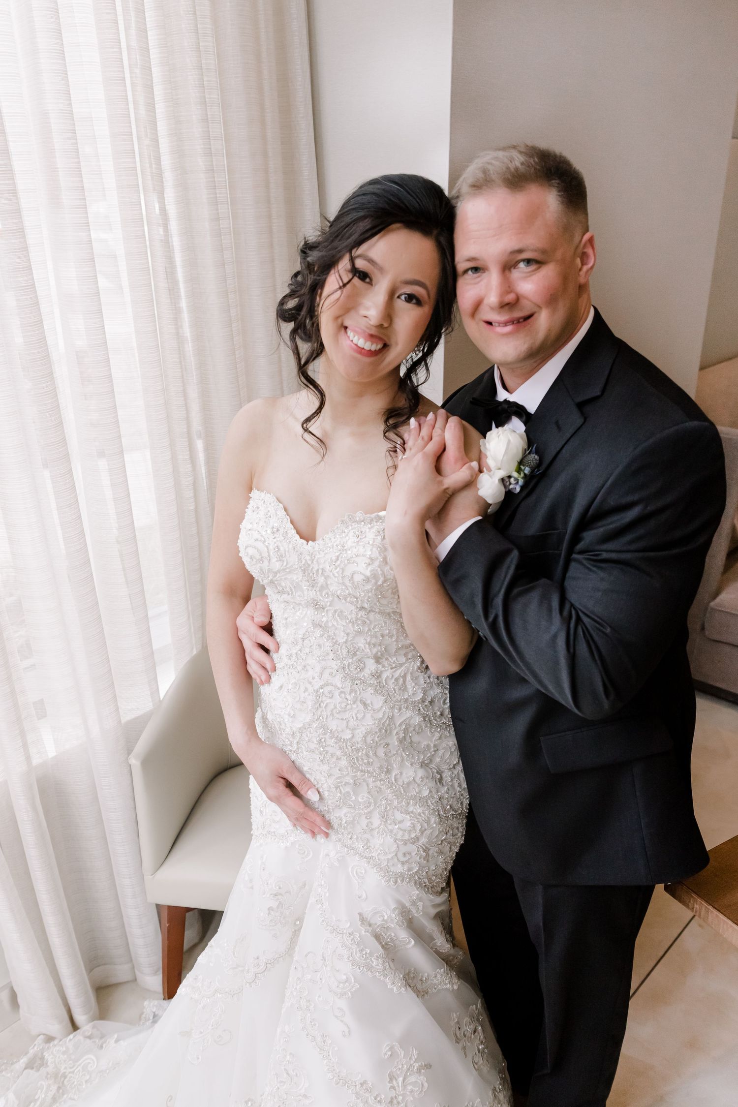A couple poses together on their wedding day in formal attire by a window.