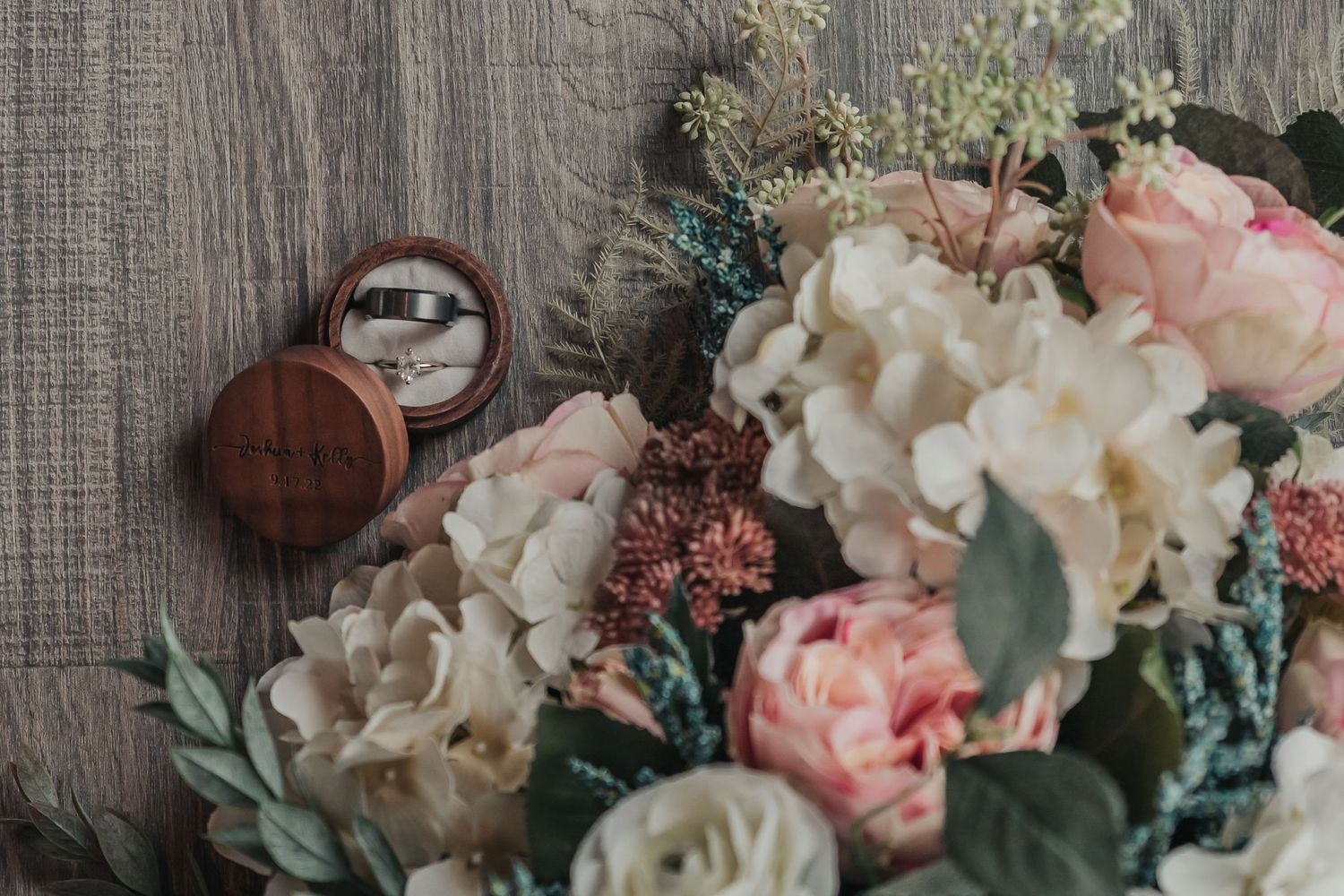 Wedding rings rest beside delicate pink and white roses with eucalyptus on a wooden surface.
