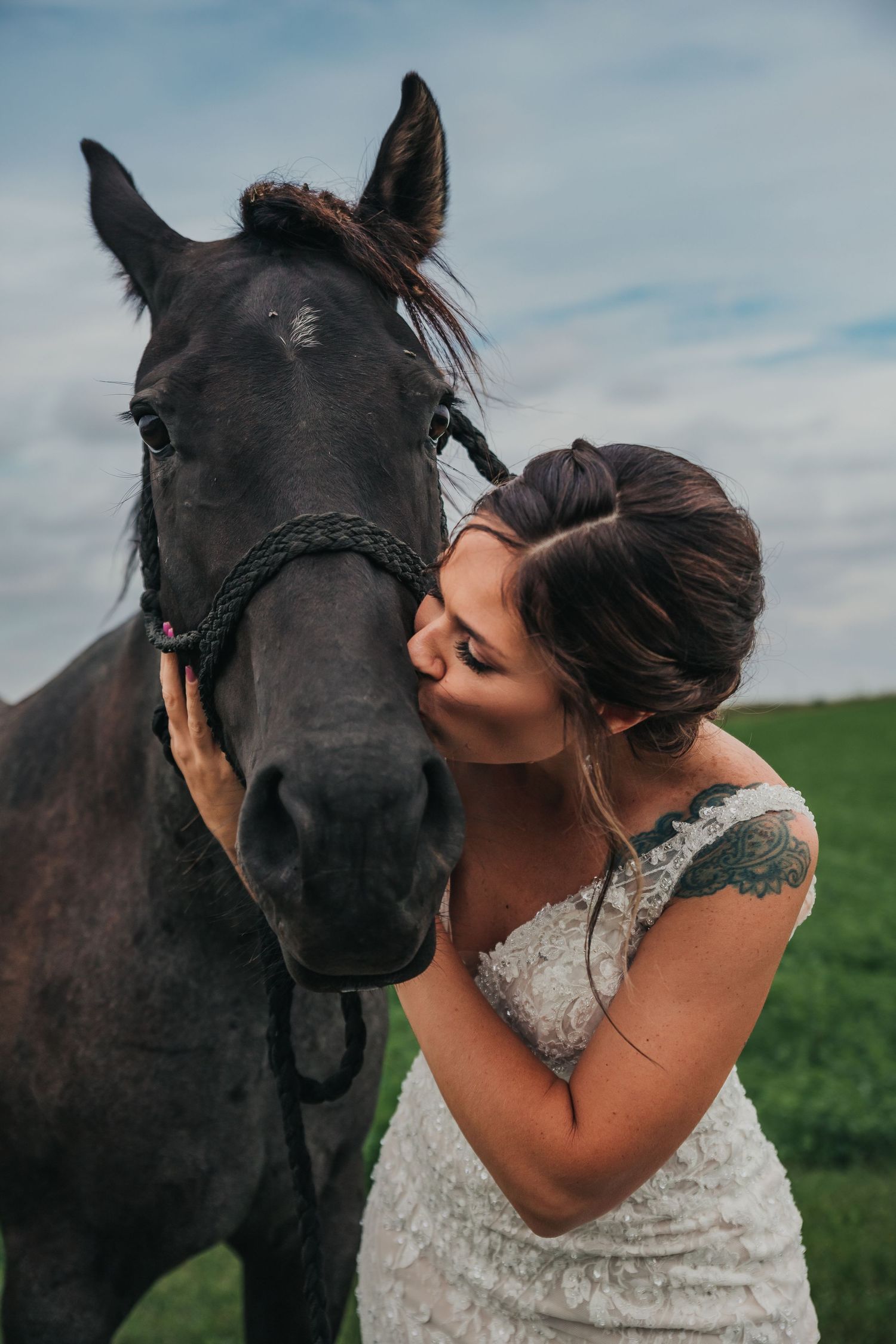 Woman in white dress sharing tender moments with a black horse in a green field.