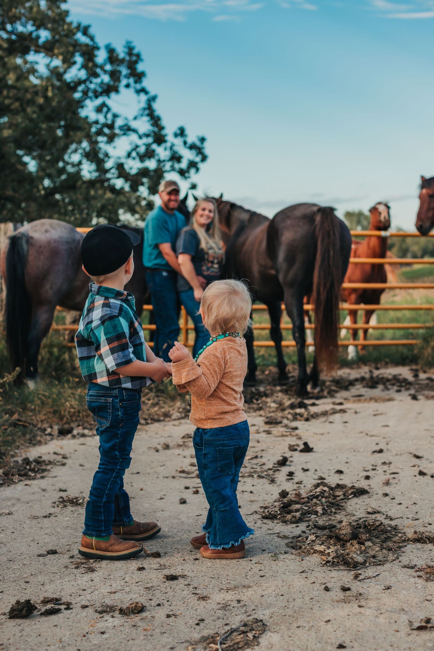 Children in western wear playing near horses in rustic ranch setting under blue sky.