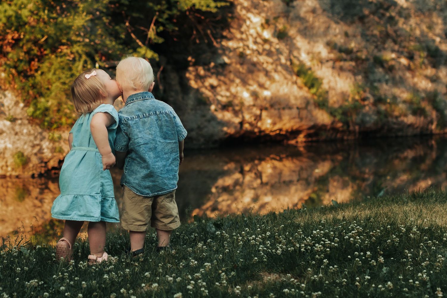 Two small children in casual clothes stand together by a pond in autumn.