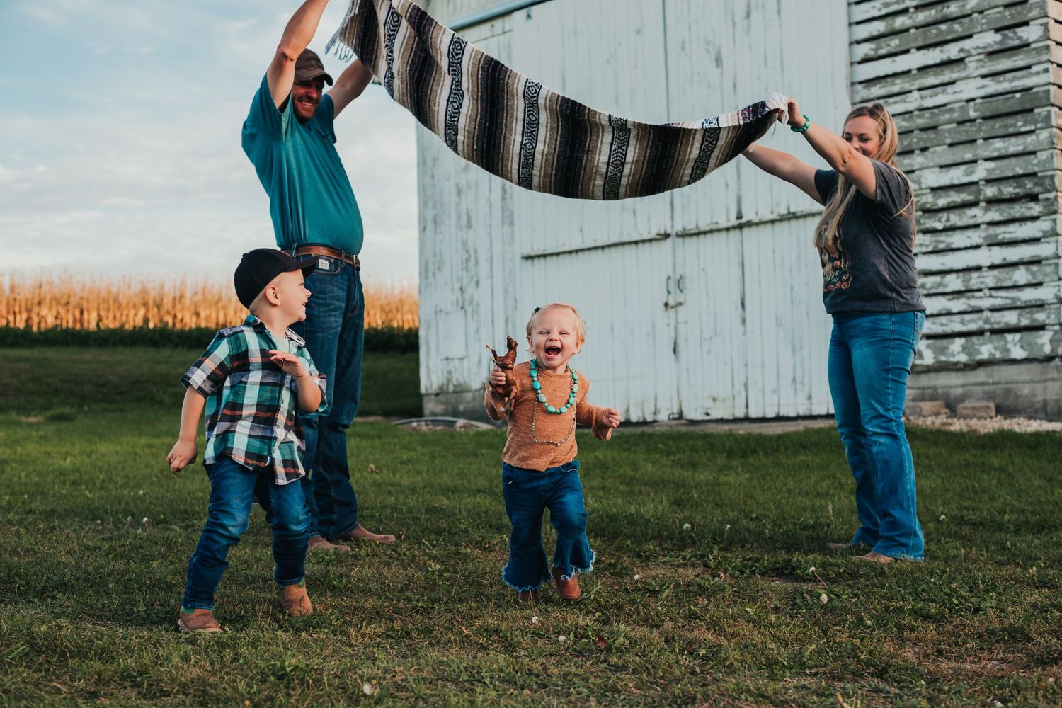 Family plays outdoors with a blanket near a white barn and cornfield at sunset.