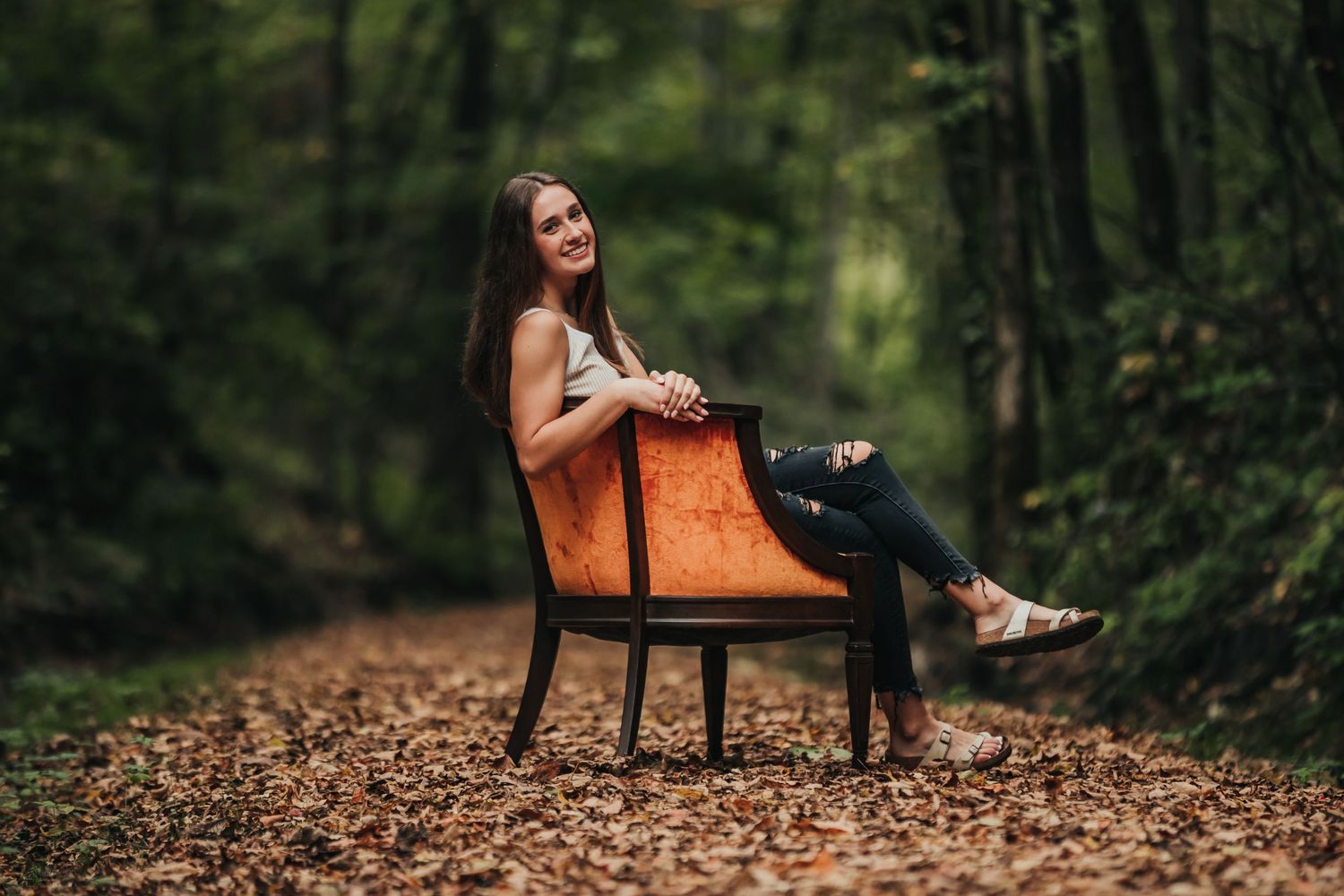 Woman in casual attire lounging on vintage leather chair in wooded forest setting.
