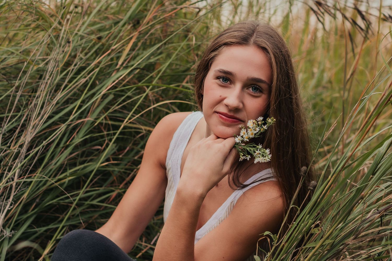 Young woman smiling with flowers in tall grass during golden hour.