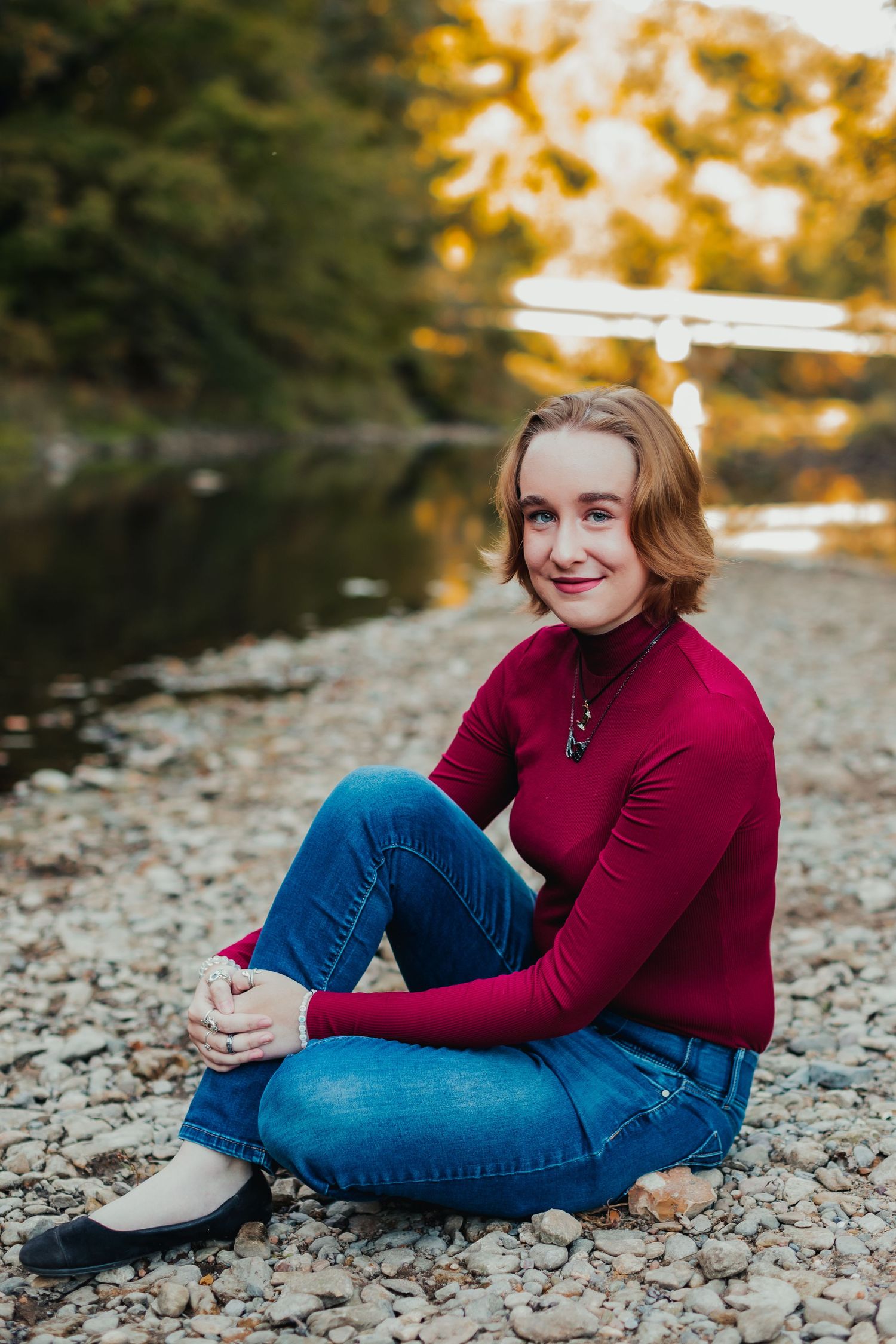 Person in burgundy sweater and jeans sitting on rocky riverbank during sunset.