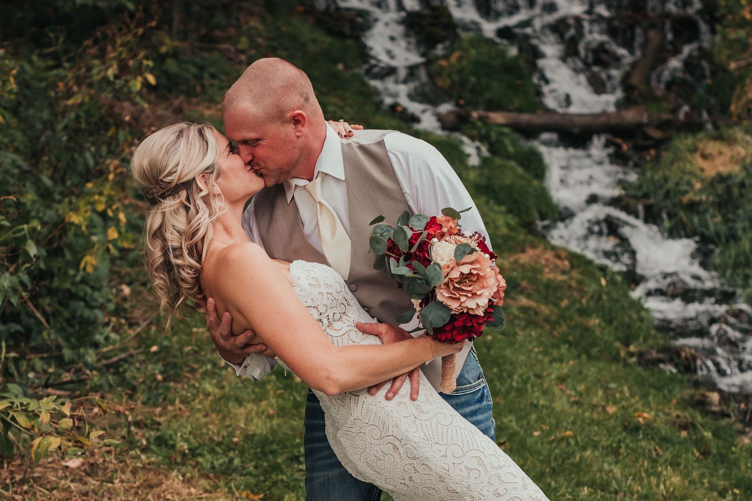 A couple shares a romantic kiss during their wedding with a waterfall in the background.